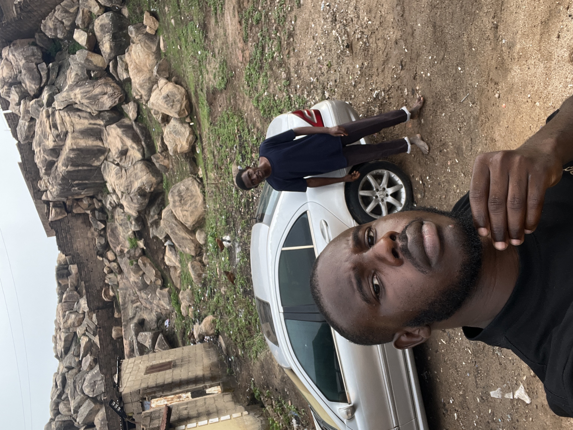 The image shows two men and a silver sedan in an outdoor setting. The man closest to the camera is taking a selfie and has a dark beard and black shirt. The second man is standing next to the car, wearing a dark shirt and pants. The car is parked on a dirt and grassy area. In the background, there is a large rock formation and a building. The location is in the city of Unknown, Unknown.