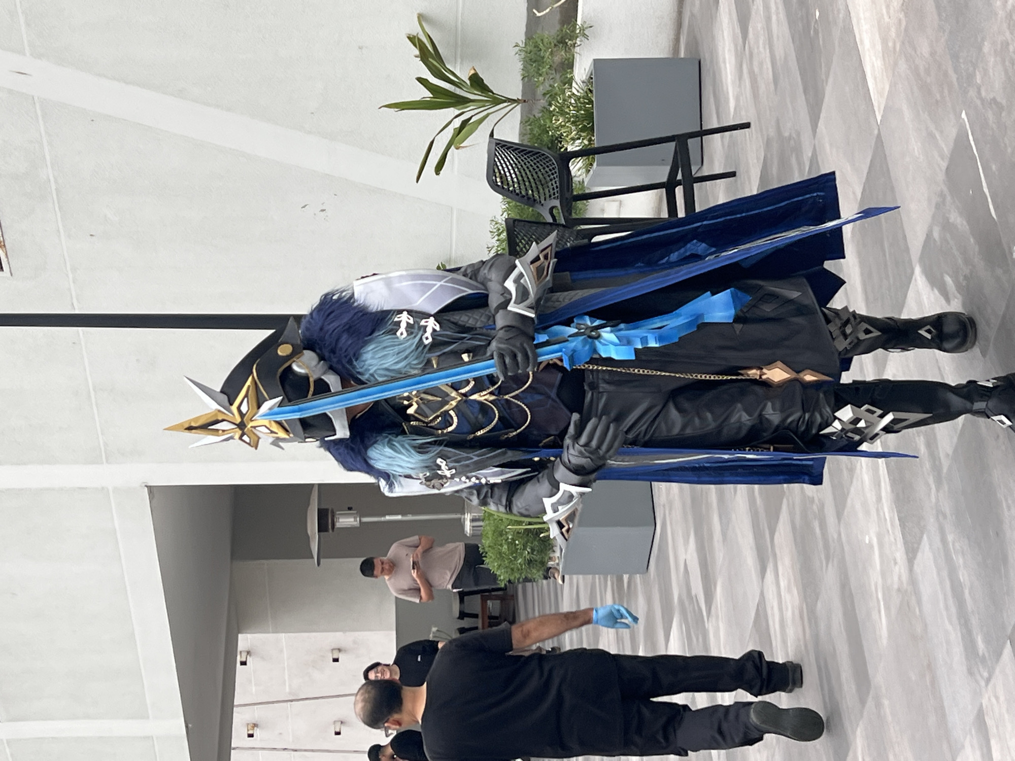 In the city of Arica, Chile, an individual in elaborate cosplay walks across an outdoor paved area. The costume features a dark, ornate helmet with gold accents, a blue fur collar, and a dark coat with a blue cape. They are holding a long, bright blue sword with a golden hilt. The individual's legs are clad in black leather pants with decorative silver elements on the thighs and boots. They are wearing black gloves. In the background, other people are visible. To the left, a person wearing a black t-shirt and pants, with light blue gloves on one hand, is walking away from the camera. Further back, another person in a light brown t-shirt and dark pants is looking at their phone. Behind them, a heat lamp stands next to a table and chairs. To the right of the cosplayer, two modern outdoor chairs with mesh seats are positioned in front of planters containing green foliage. The environment appears to be an urban outdoor space, possibly a plaza or courtyard, with a light-colored wall as a backdrop. The paving stones of the ground are laid in a geometric pattern.