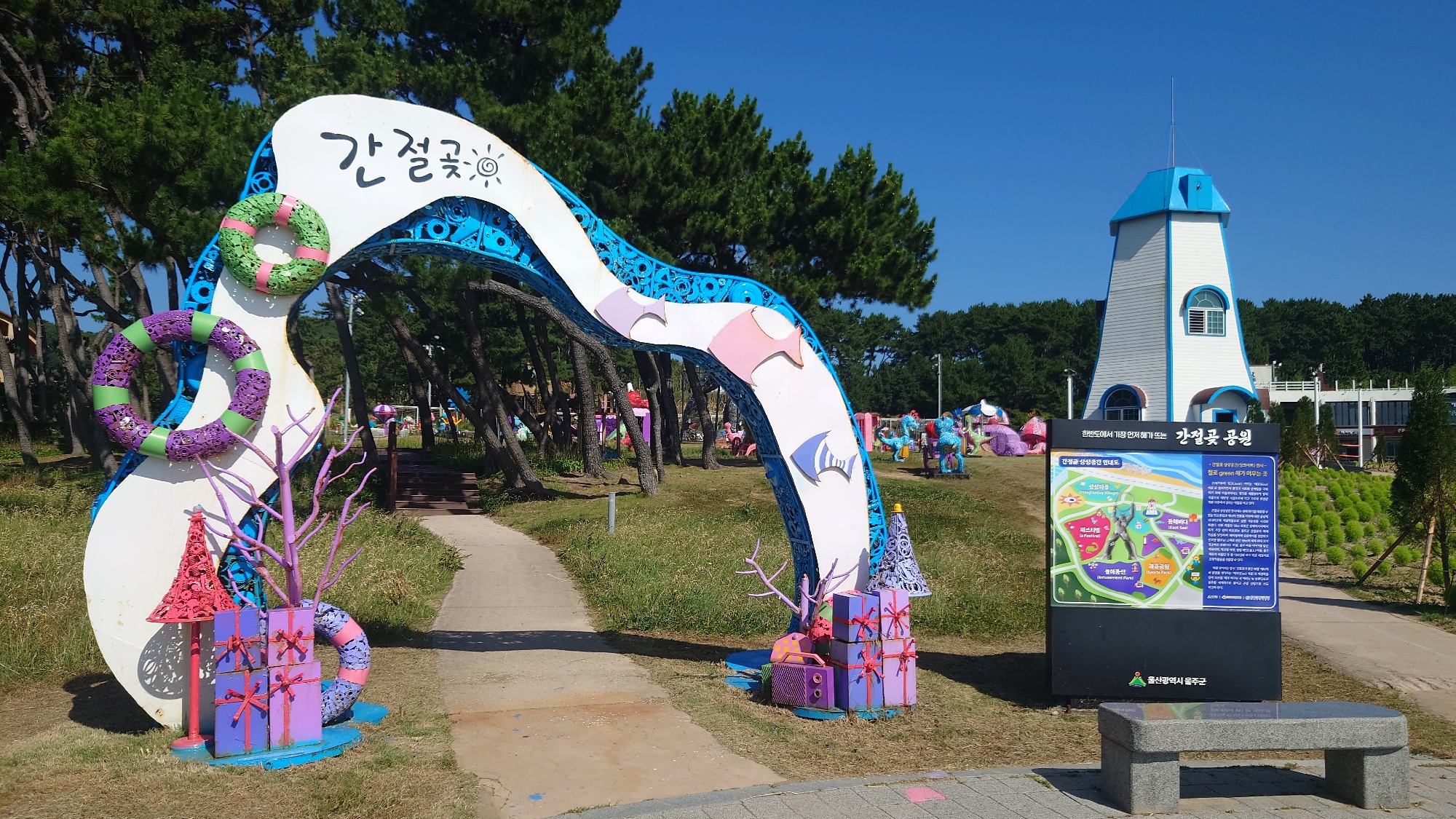 The image shows an outdoor scene at a park in Ulsan, South Korea. In the foreground, a large, white and blue arched entryway decorated with colorful buoys and gift boxes stands on a grassy area. To the right of the arch is a large informational sign with a map of the park and text in Korean, placed next to a stone bench. Behind the sign is a white lighthouse-like building with a blue roof, surrounded by trees and some decorative sculptures. The sky is clear and blue, indicating a sunny day. The overall atmosphere is cheerful and welcoming, suggesting a public park or recreational area. The sign identifies the location as "Gangjeolgot Park" (강절곶 공원).