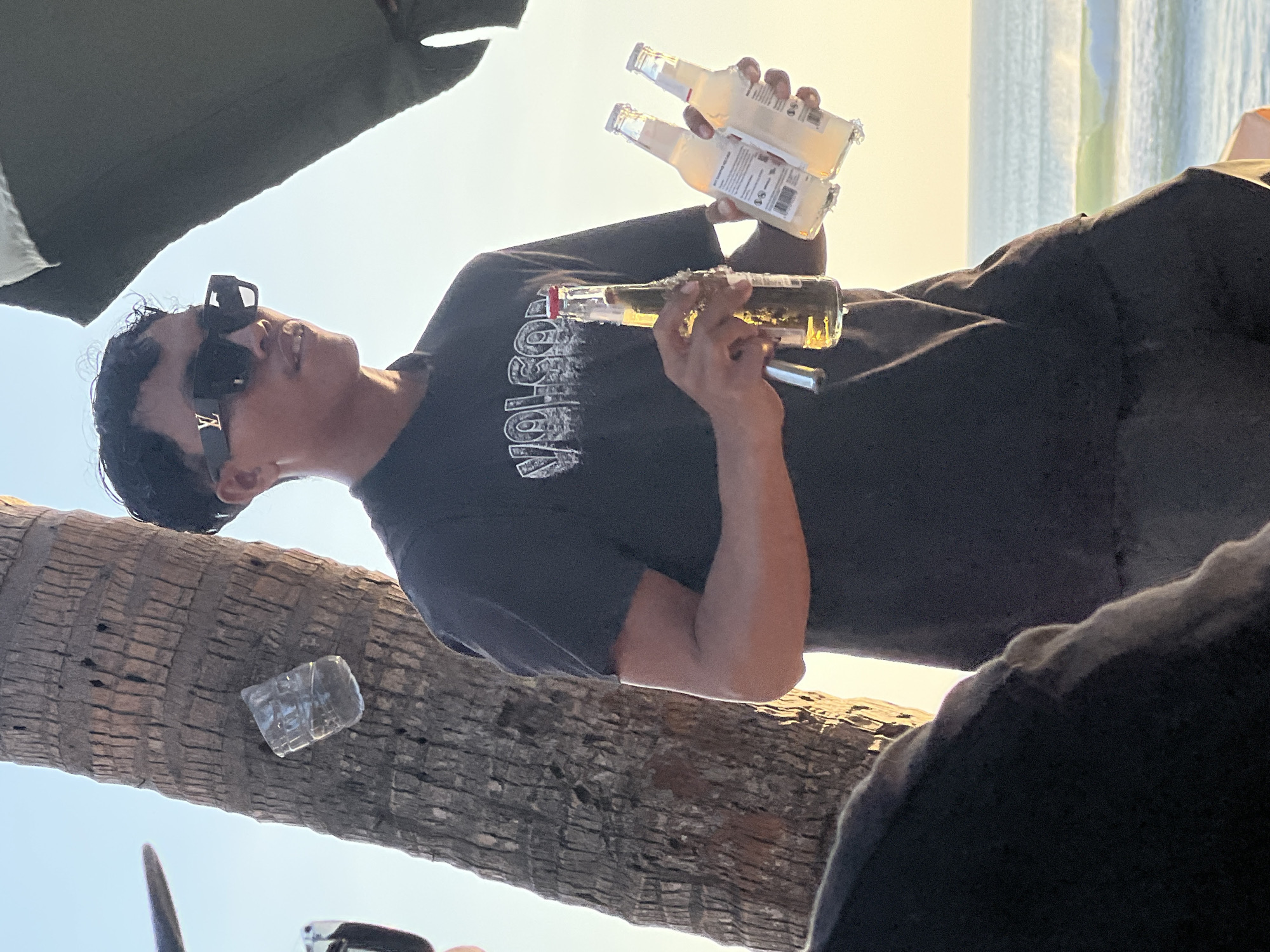 A young man stands near a palm tree by the ocean, holding two bottles of a light-colored beverage and one bottle of beer. He wears a black t-shirt with a "VOLCOM" logo and black sunglasses with a gold "LV" emblem. A plastic cup is attached to the palm tree. The background features a calm ocean with small waves and a bright sky, suggesting a sunset or sunrise. The overall setting appears to be a beach in Kuta, Indonesia.