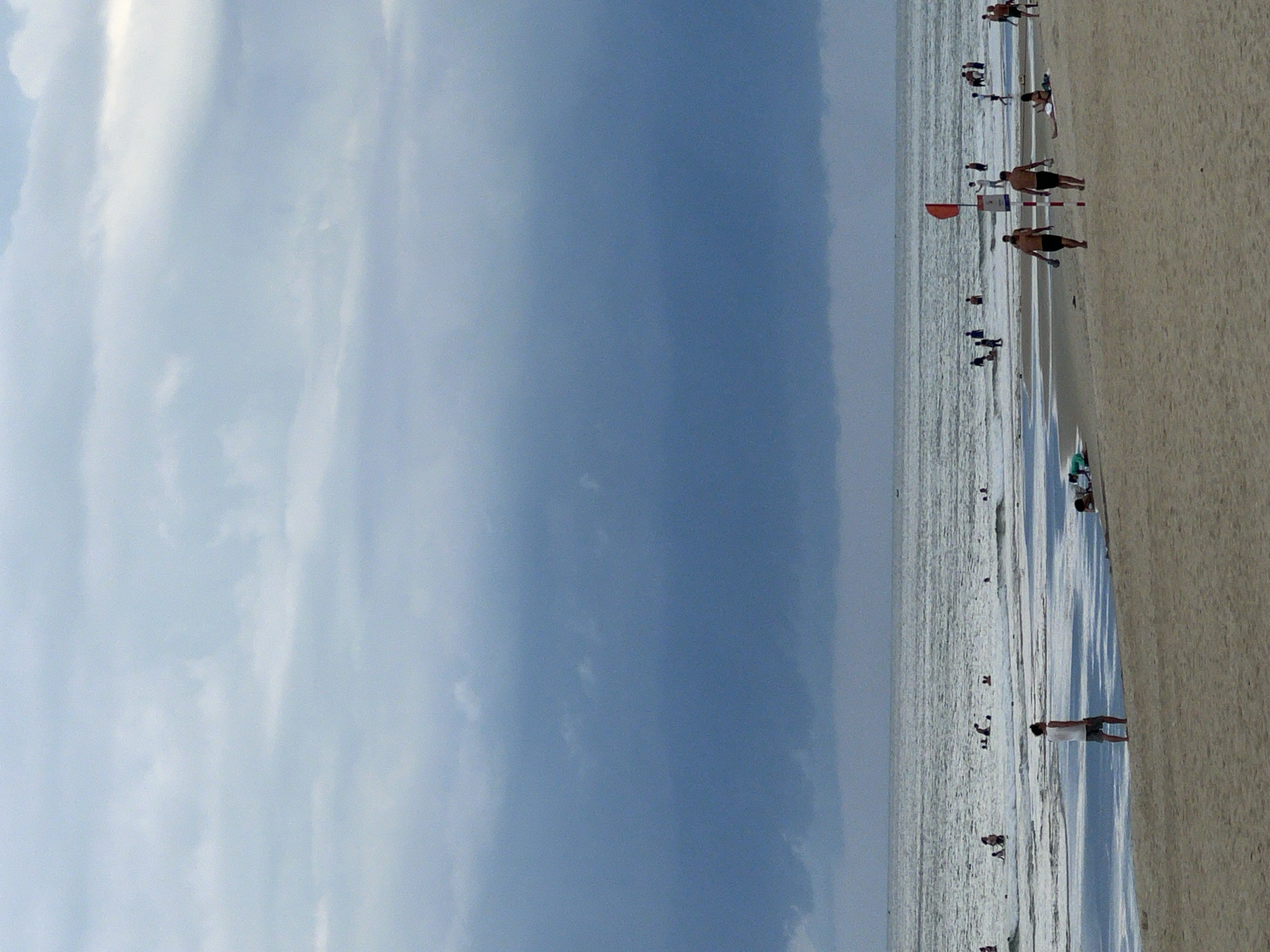 This is a photograph of a beach scene in Đà Nẵng, Vietnam, rotated 90 degrees clockwise. The image is dominated by a vast expanse of pale blue sky filled with scattered white clouds. Below the sky, the ocean stretches out, appearing calm with gentle waves. A sandy beach occupies the right side of the frame, with footprints visible on the damp sand near the water's edge.
Several people are scattered across the beach and in the shallow water. Some are standing, while others appear to be walking or engaging in various activities, though the distance and resolution make specific actions difficult to discern. In the foreground on the beach, two men in dark shorts are visible, facing away from the camera. Further down the beach, near the water, a person in a white shirt and dark shorts stands. A red flag is planted in the sand near the water, a common sight on beaches for signaling conditions.
The lighting suggests it is daytime, likely mid-morning or afternoon, with the sun casting soft shadows. The weather appears to be clear and pleasant for a beach outing. The overall mood is one of leisure and relaxation, characteristic of a day at the seaside. No text is visible in the image.