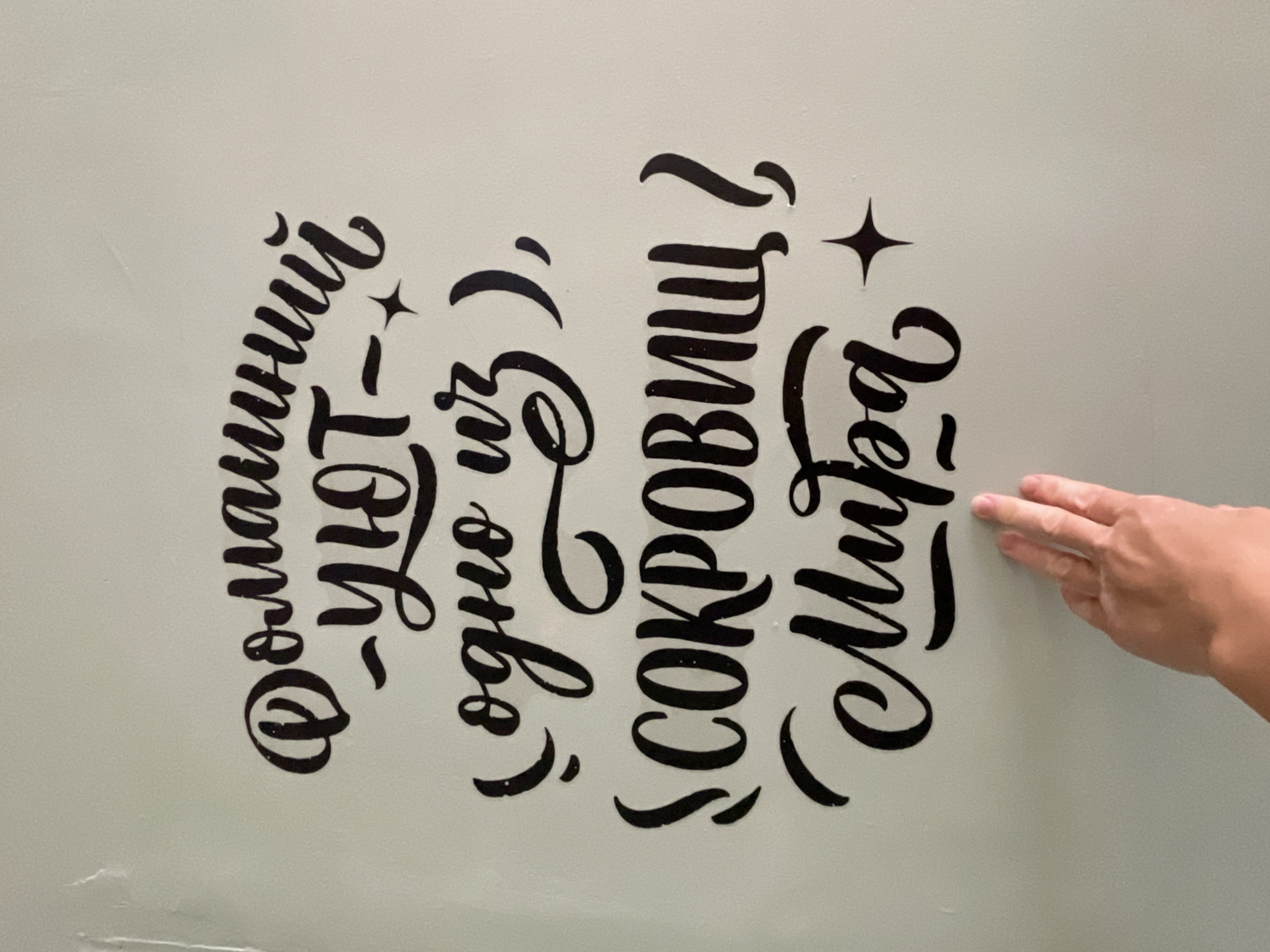The image shows black Cyrillic text on a light greenish-grey wall, likely a painted surface. The text, in a stylized font with flourishes, reads "Романтика - одно из сокровищ Мира?" It includes stylized star elements. A human hand is visible to the right of the text, with two fingers pointing toward the word "Мира". The location, Sochi, Russia, is not evident from the image itself but can be inferred if that information is part of the request context.