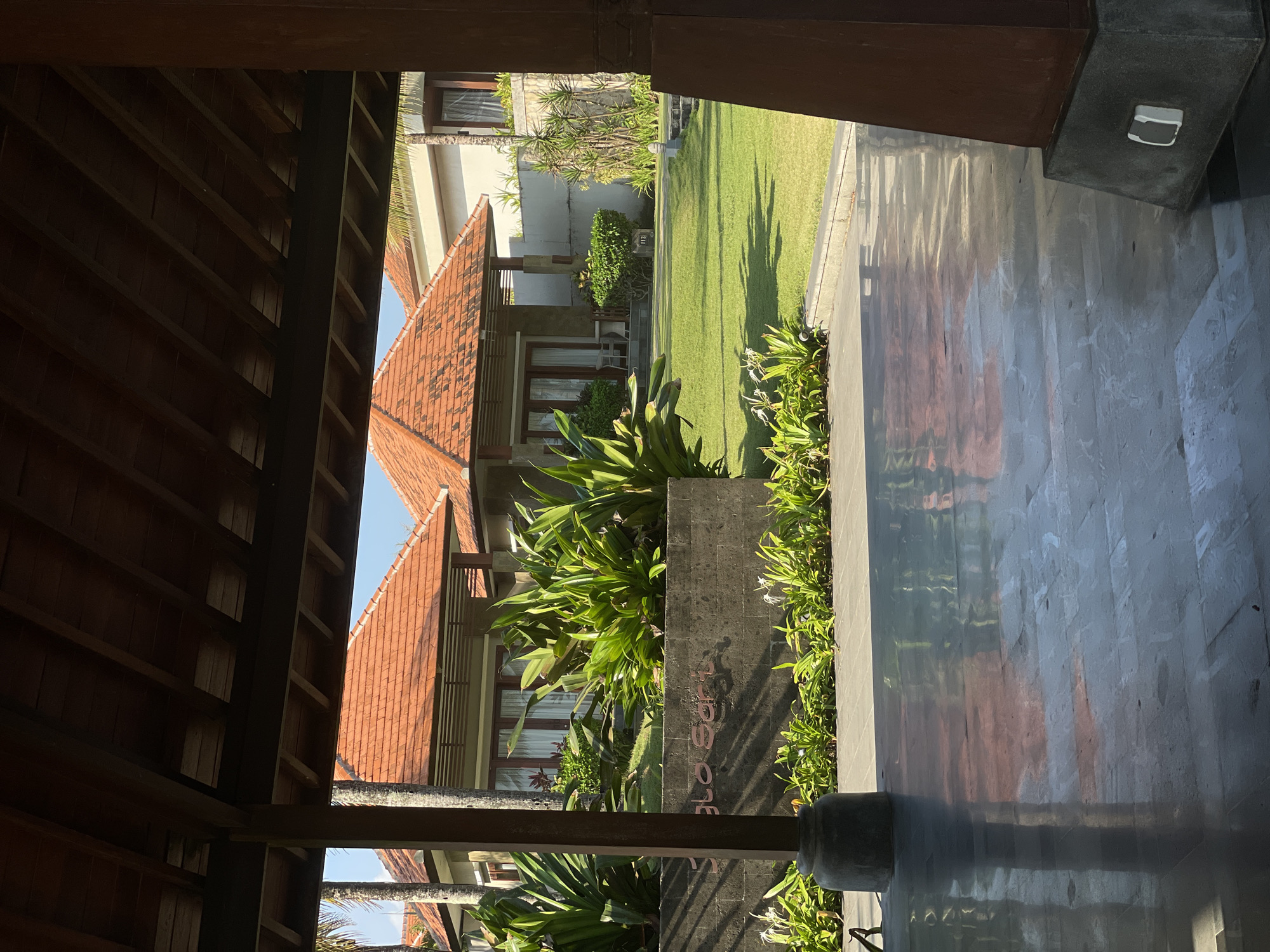 The image shows a resort setting in Selemadeg Timur, Indonesia. In the foreground, a polished dark tiled floor reflects the wooden roof structure above. To the left, a substantial concrete pillar supports the roof. Beyond the foreground, a manicured green lawn stretches towards buildings with red tile roofs. A low, dark grey stone wall separates the tiled area from the lawn. Lush green plants, including large-leafed tropical foliage, are planted behind the wall and on the lawn. In the background, the buildings appear to be villas or resort rooms, with visible windows and doors. The overall scene conveys a serene and tropical ambiance. There are no people visible in the image.
