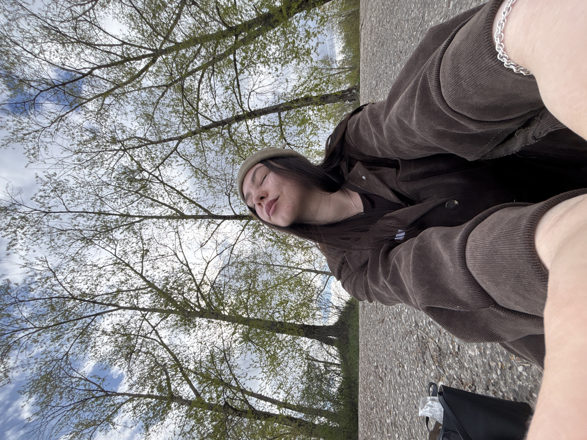 A young woman with long, dark hair, wearing a brown corduroy jacket, a dark top, and a beige beret, sits on a gray paved surface with a black bag nearby. She has her eyes closed and appears to be looking up at trees with sparse green leaves against a cloudy sky with patches of blue. The photograph is taken from a low angle, with the trees in the background. Her left wrist is visible, adorned with a silver chain bracelet. The paved surface extends into the distance, suggesting a park or similar outdoor location. Based on the vegetation and climate, it is plausible the image was taken in a city like Perm, Russia, given the prevalence of deciduous trees and a temperate climate.