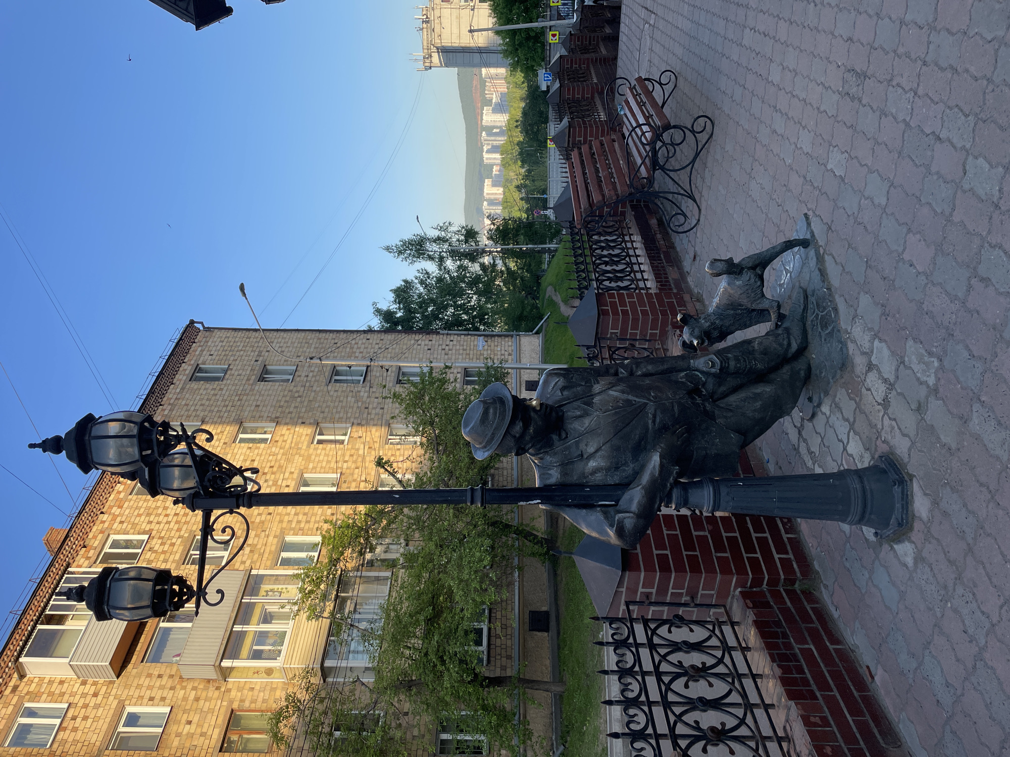 The image captures an urban landscape in Krasnoyarsk, Russia, under a clear blue sky. The primary subject is a bronze statue depicting a man in a fedora hat and a long coat, leaning contemplatively against a decorative black lamppost with multiple ornate lanterns. At his feet, a small dog looks up towards him.
Adjacent to the statue, a park bench with dark wooden slats and elegant wrought iron supports is situated along a low red brick wall, which is topped with a black wrought iron fence. The ground in the foreground consists of a patterned paved walkway made of grey and reddish-brown interlocking bricks.
In the mid-ground, a multi-story residential building with light-colored brickwork, featuring numerous windows and balconies, stands behind the lamppost and statue. Various green trees and foliage are visible around the building and behind the brick wall.
The background offers a panoramic view of a distant cityscape, with buildings nestled against a backdrop of rolling, tree-covered hills or mountains. The lighting suggests either late afternoon or early morning, with warm sunlight illuminating the scene and casting long shadows on the paved ground near the statue and bench. Overhead wires are visible against the sky, where a small bird can also be seen soaring. The scene is static, with no human activity, focusing instead on the contemplative nature of the statue within its urban setting. No visible text is discernible.