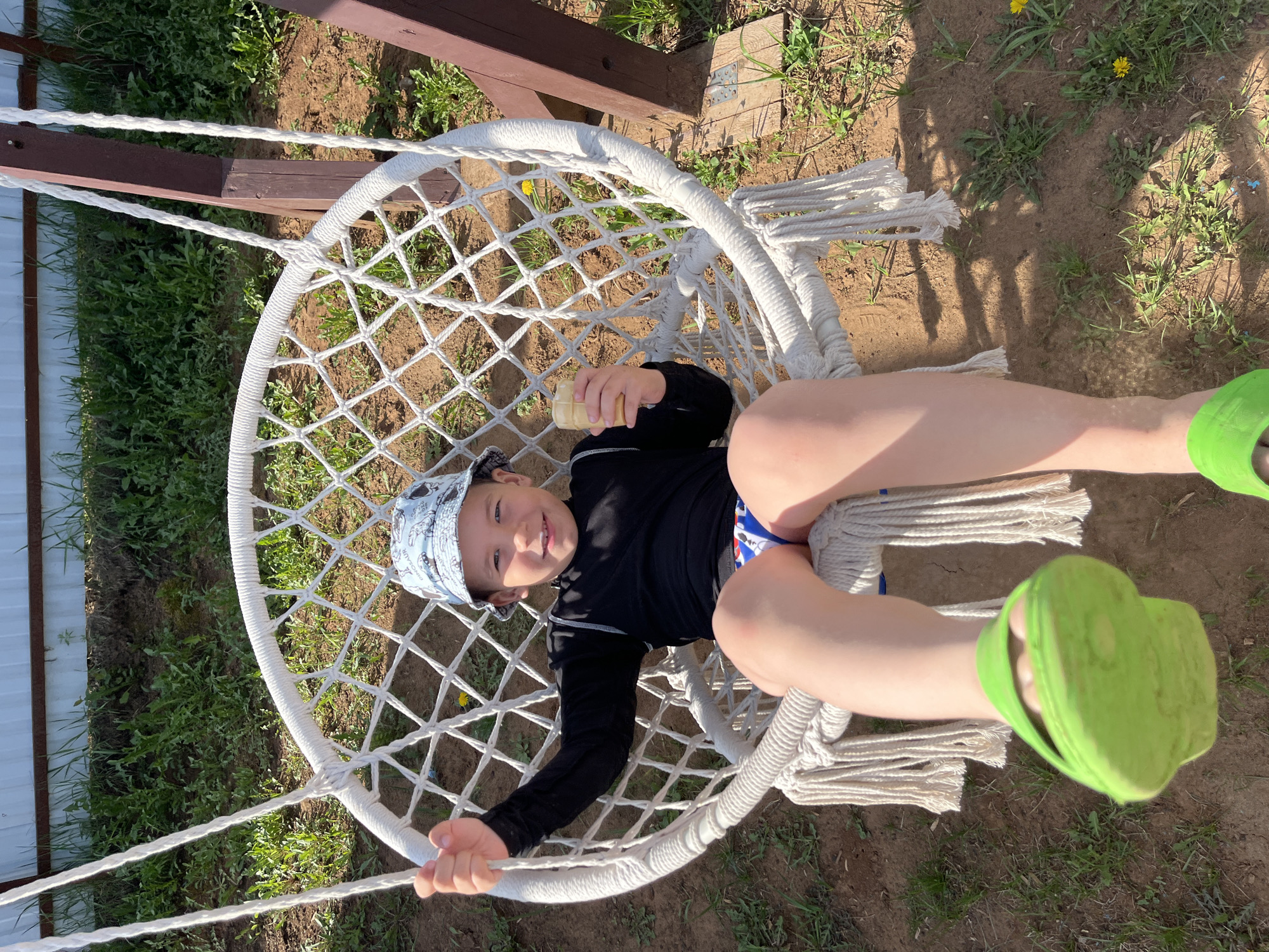 A young child is lying in a white macrame swing chair in an outdoor setting. The child is wearing a blue patterned hat, a black long-sleeved shirt, and swim trunks. They are also wearing bright green clogs. The child is holding a small, yellow container in one hand and is smiling at the camera. The swing is suspended from a wooden frame. The ground is a mix of grass and dirt, with some dandelions visible. The location appears to be a yard or garden. The city and country are Chaikovsky, Russia.