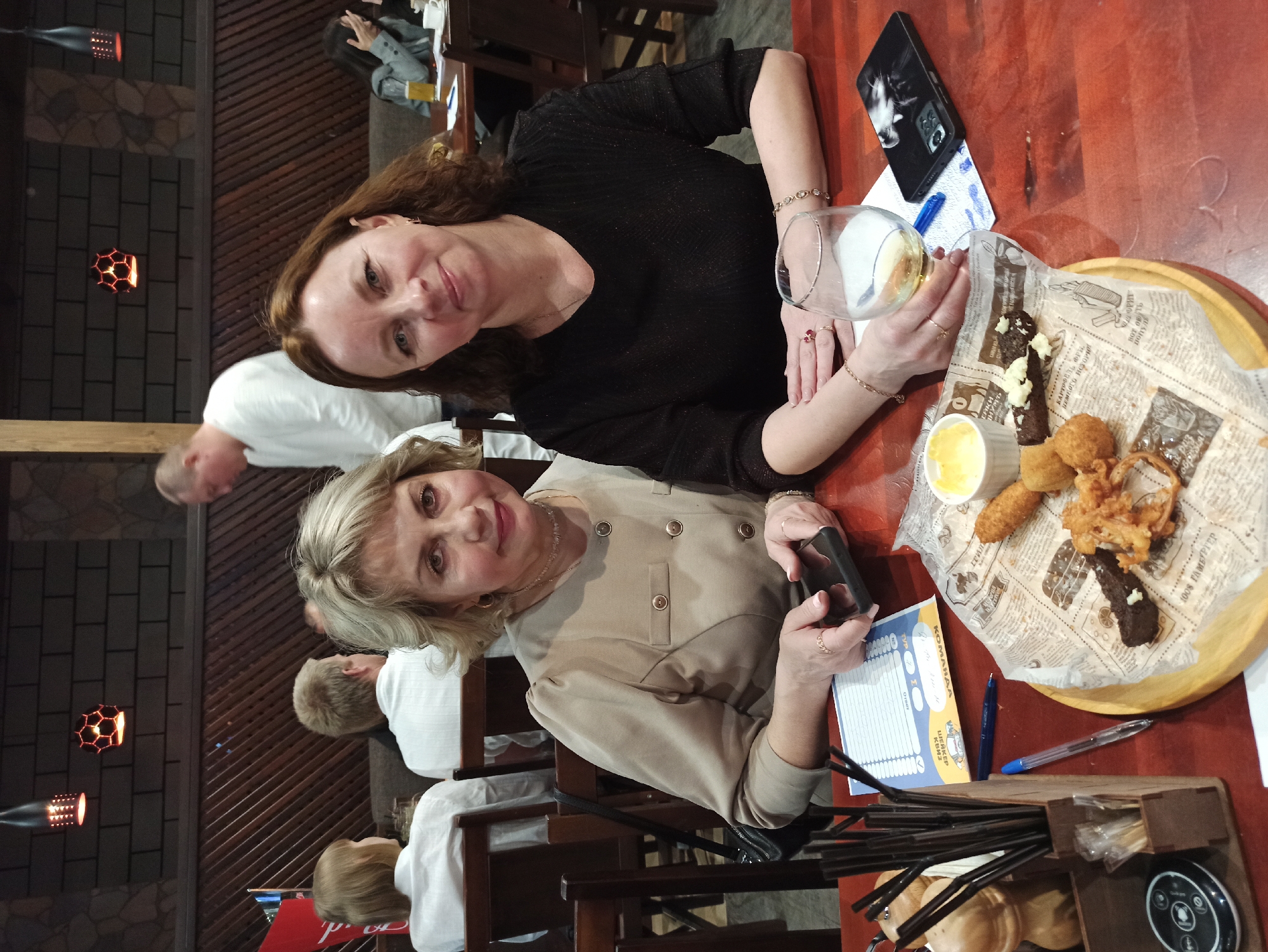 Two women are seated at a wooden table in what appears to be a dimly lit restaurant or bar. The image is oriented sideways, with the subjects looking towards the camera. The woman in the foreground has blonde hair and is wearing a light-colored, buttoned jacket, holding a black smartphone in her hands. The woman behind her has brown hair, wears a black top, and holds a glass containing a drink, possibly white wine. Both women have subtle smiles.
On the table in front of them, a wooden tray holds various fried snacks, including what resembles onion rings, corn dogs, and dark bread with a white topping, all served on paper resembling newspaper. A small white bowl with a yellow dip is also on the tray. Other items on the table include a black smartphone with a cat-themed case, a pen, and a score sheet or game card. A wooden caddy filled with straws and other utensils is also visible.
In the background, other patrons are out of focus, and the decor includes dark wooden paneling and decorative light fixtures. The visible text on the game card includes "КОМАНДА" (Team), "Ответ" (Answer), and "ШЕЙКЕР". Text on the newspaper wrapping mentions "КАРТОФЕЛЬ ФРИ" (French fries), "НЕМНОГО ИСТОРИЯ" (A little history), and "100% ГАМБУРГЕР" (100% Hamburger). The setting suggests a casual social gathering, possibly a quiz night or a meal with friends.