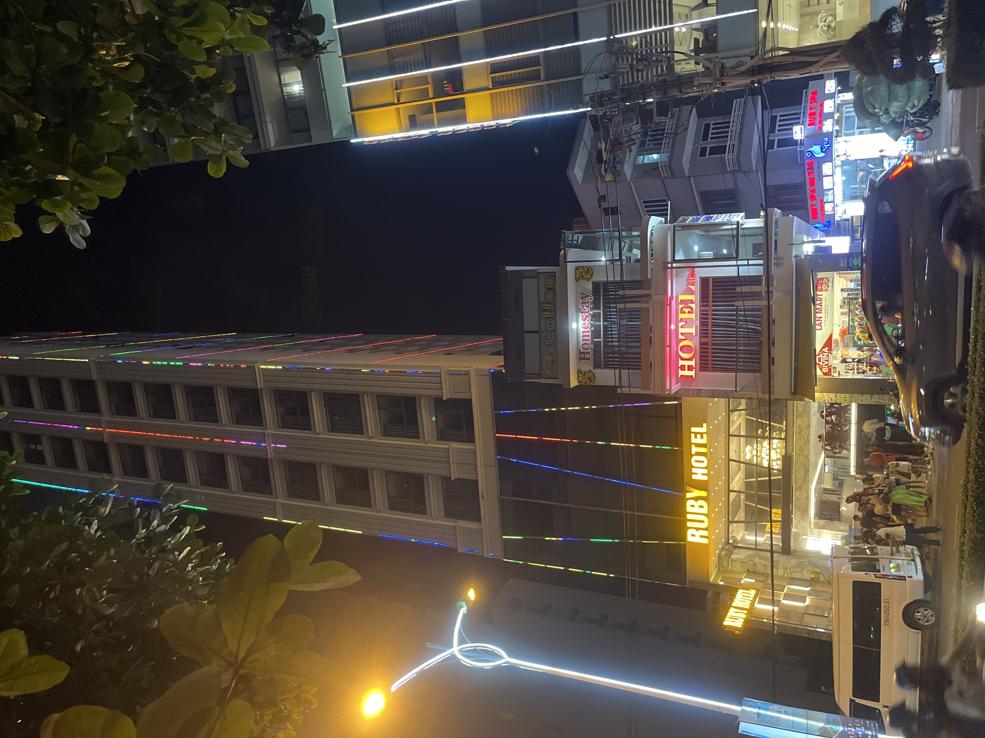 The image depicts a nighttime street scene in Nha Trang, Vietnam. A tall, modern building, identified as "RUBY HOTEL," is prominently featured, adorned with vertical strings of colorful lights. Adjacent to it, another building displays bright white vertical lights. In the foreground, a white van with the license plate "79H-050.91" is parked on the side of the street, and a group of people are gathered nearby. A dark-colored SUV is also visible in the street. Other businesses, including "Coffee" and "Homestay," are present on the lower floors of the buildings. The street is illuminated by streetlights and the vibrant building lights, creating a lively urban atmosphere. Trees with green foliage are visible in the upper portions of the image, contrasting with the dark night sky.