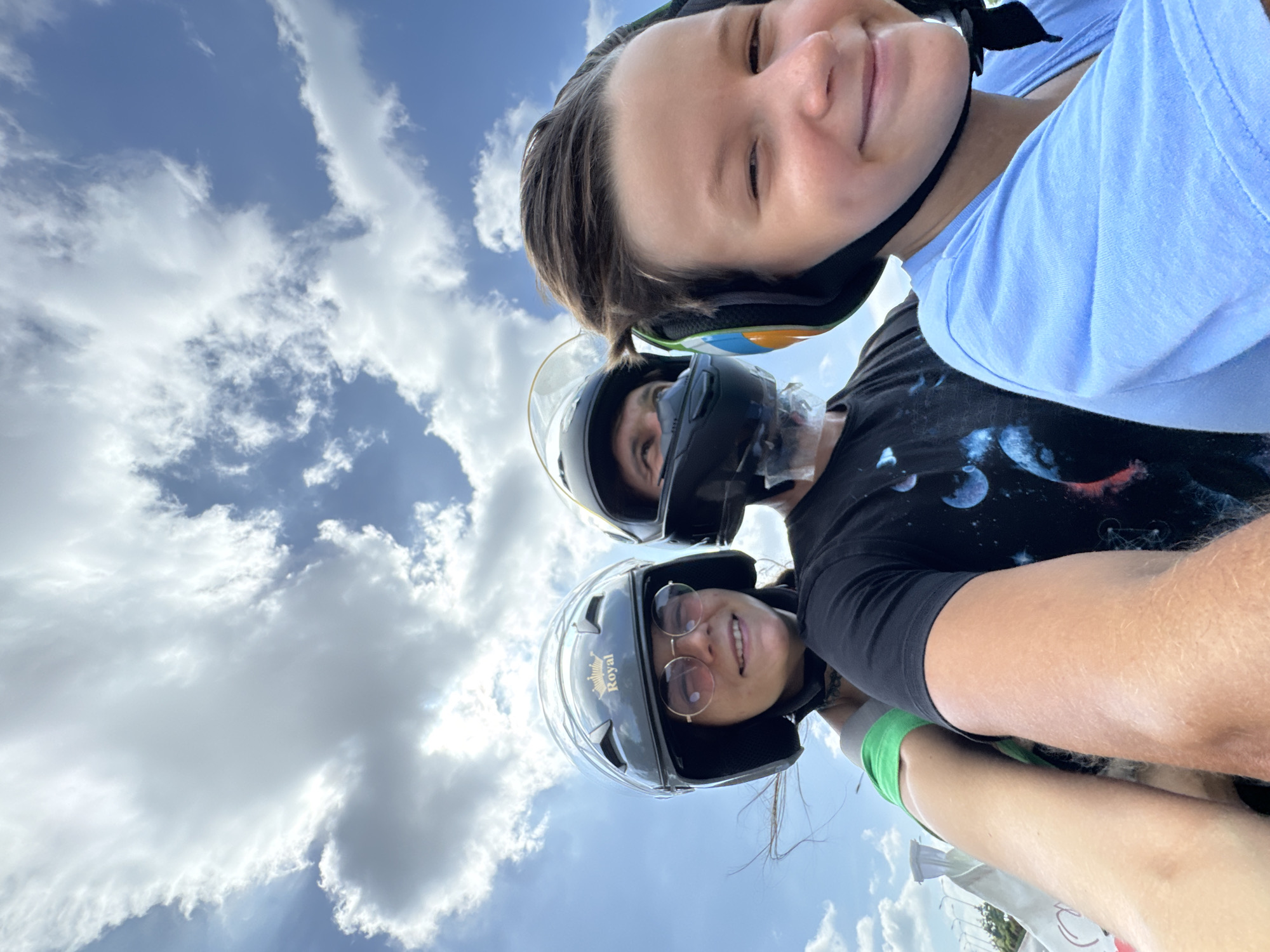 The image depicts three individuals, presumably on a motorcycle or scooter, captured in a low-angle selfie against a bright blue sky with scattered white clouds. The scene is located in Mui Ne, Vietnam.
In the immediate foreground, on the right, is a young person, possibly male, with dark hair, wearing a multi-colored open-face helmet (green, orange, blue, black) and a light blue short-sleeved t-shirt. This individual is smiling directly at the camera.
Behind and slightly to the left of the foreground subject is an adult male, visible from the neck up, wearing a black t-shirt with a blue and purple cosmic graphic. He wears a black full-face helmet with the clear visor lifted, revealing a clear plastic chin shield or mask below his chin. His gaze is directed slightly upwards and to the left. His right arm, tan and hairy, is partially visible.
Further back on the left is an adult female, smiling and looking towards the left. She wears a dark grey open-face helmet with a gold "Royal" brand crown logo on the front. She has round, rose-tinted sunglasses on and is wearing a green top or strap. Strands of her dark hair are visible, blown by the wind. A white bag with red markings is partially visible near her left arm.
The sky occupies the majority of the upper frame, showcasing clear blue areas interspersed with various white cumulus and cirrus clouds. The lower left portion of the image subtly reveals structures consistent with a roadside environment, including faint light poles and a hint of a road. The wind-blown hair of the subjects suggests motion.