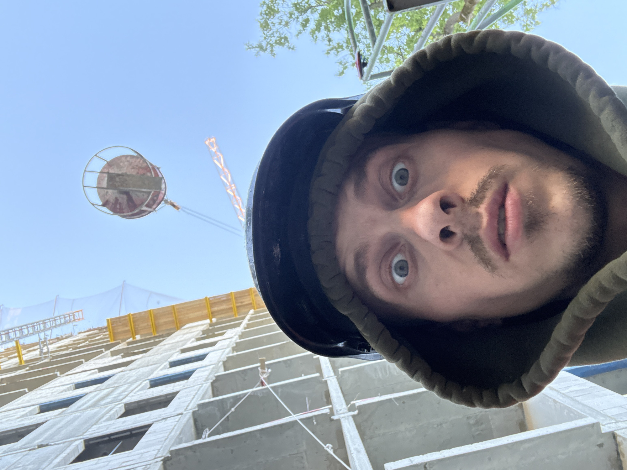 A man wearing a black hard hat and a dark green hooded jacket takes a selfie outdoors. He has light brown hair, blue eyes, a mustache, and a goatee. His mouth is slightly open, and his eyes are wide with surprise. In the background, a tall building under construction is visible, with scaffolding and netting. A crane is also present, with a circular object hanging from it. The sky is clear and blue. The overall scene suggests a construction site. The image context does not provide any information about the location being Moscow, Russia.