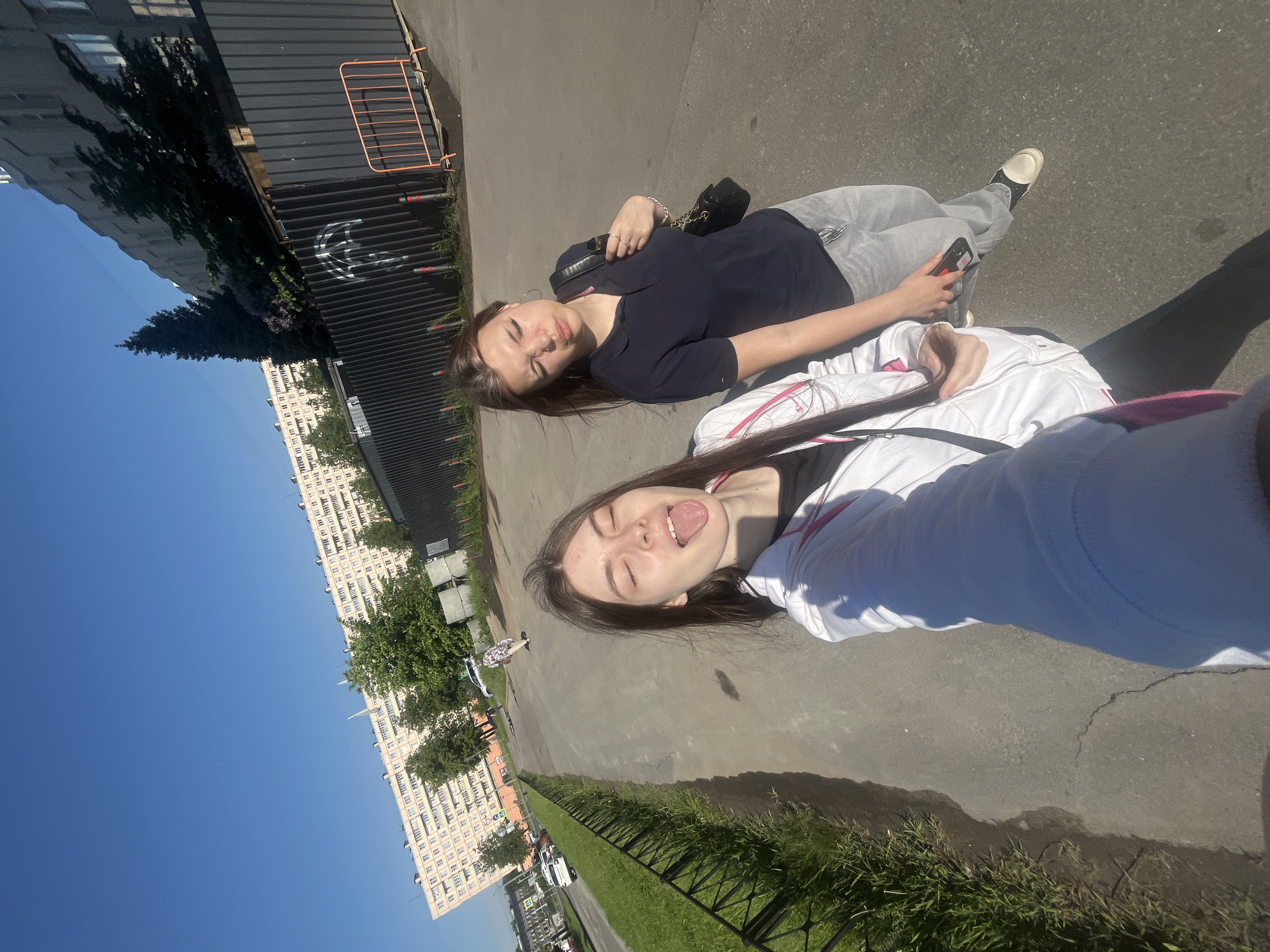 The image displays two young women posing for a photo outdoors under a clear blue sky, with the entire scene rotated 90 degrees clockwise. They appear to be lying on a paved surface, looking up towards the camera.
The woman on the left has long, dark hair and is dressed in a dark blue polo shirt, light grey pants, and light-colored shoes. She wears a black shoulder bag, silver hoop earrings, a nose piercing, and holds a red-cased smartphone in her right hand. Her expression is relaxed, with her eyes slightly narrowed against the bright sunlight.
The woman on the right, who seems to be taking the photo, also has long, dark hair and is wearing a white and black hoodie. She is playfully sticking her tongue out, with her eyes closed, suggesting a lighthearted mood.
In the background, a large, multi-story apartment building with numerous windows extends across the horizon, indicating an urban or suburban setting. To the immediate left of the women, there is a dark, corrugated metal fence adorned with a white graffiti symbol resembling a stylized crescent or 'C'. An orange construction barrier is visible behind this fence. Lush green trees and bushes line the paved path, with bright green grass extending further back. The strong shadows cast by the bright sunlight suggest it is midday or early afternoon. No legible text is visible on clothing or signs.