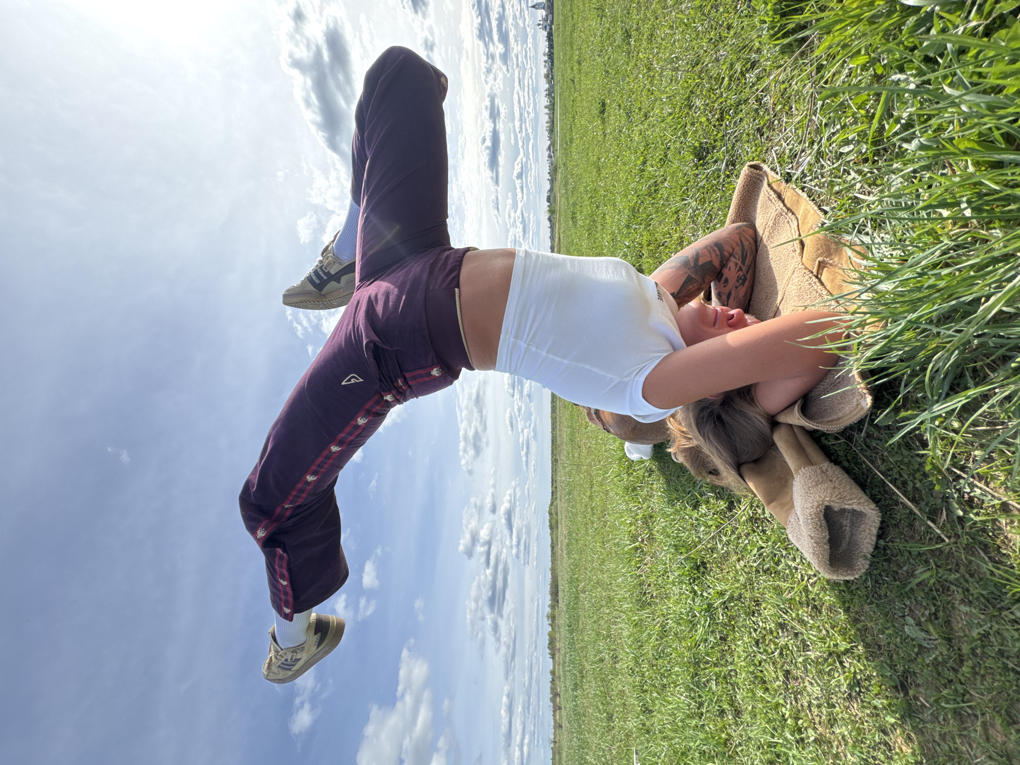 A person is performing a backbend in a grassy field under a partly cloudy sky. The person is wearing a white t-shirt, maroon track pants with a patterned stripe, and sneakers. The person is lying on a tan blanket. The location is Bol'shoye Kozino, Russia.