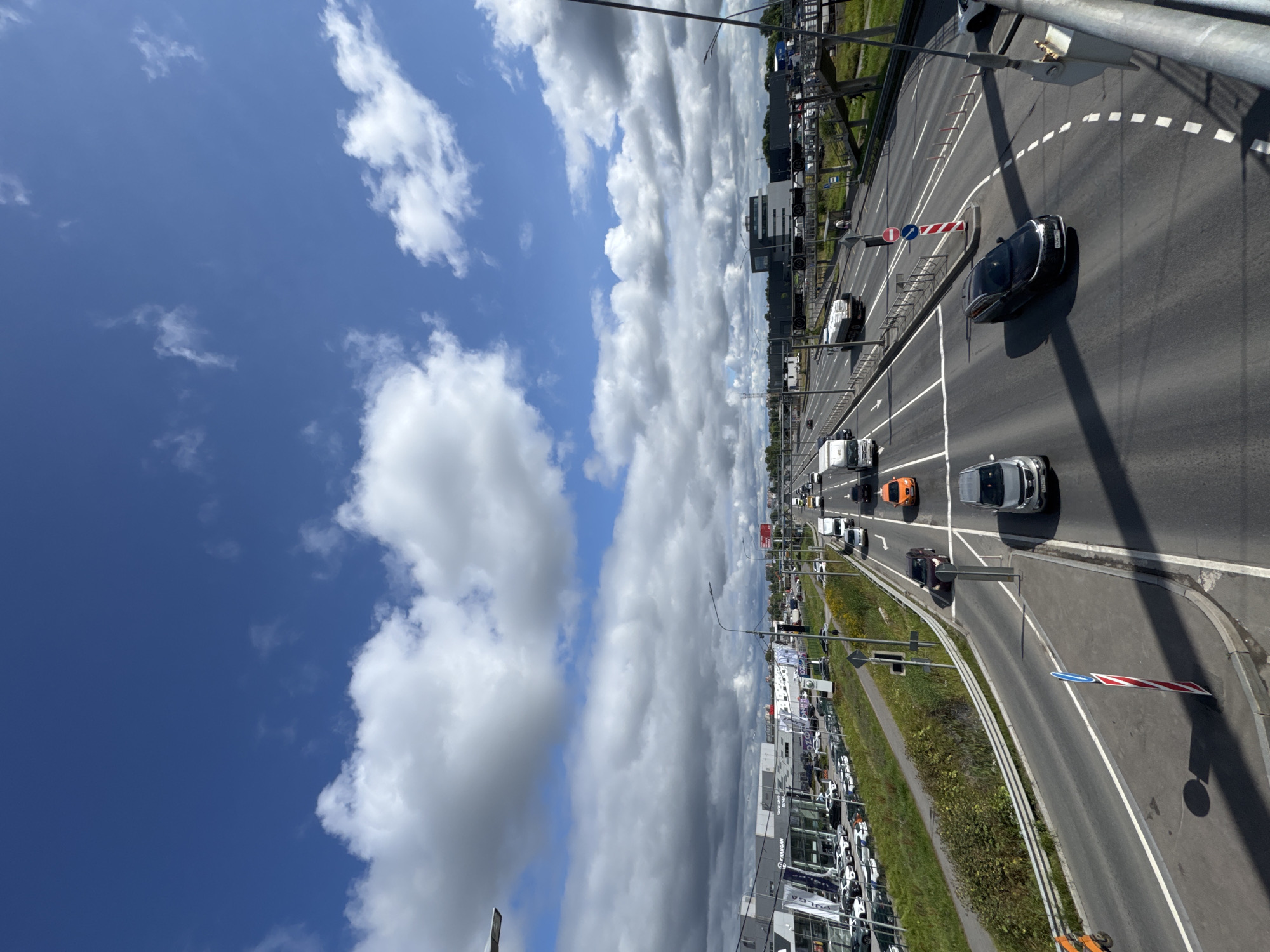 The image shows a view of a highway in Saint Petersburg, Russia, under a blue sky with scattered clouds. The highway is multi-lane and carries a moderate amount of traffic, including cars, vans, and trucks. On the left side of the highway, there are several car dealerships with numerous vehicles parked on their lots. Buildings of commercial and industrial nature are visible in the background. On the right side of the highway, there are pedestrian overpasses and some greenery. The perspective is from an elevated viewpoint, likely a bridge or overpass, looking down at the road.