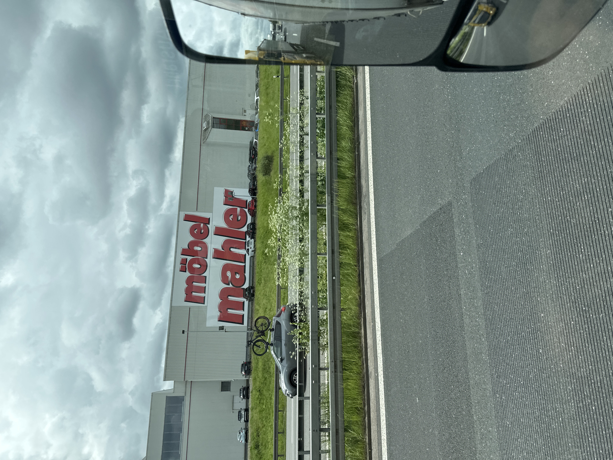 The image shows a large building with the prominent red lettering "möbel mahler" on its facade. The building is a furniture store, commonly referred to as "Möbel Mahler Nossen," located in Nossen, Germany. In front of the building, there is a highway with a grey car driving on it. The car has a bicycle mounted on its roof. There are other cars parked in the background. The sky is overcast with clouds. The photo appears to be taken from another vehicle on the highway, likely a truck, as a side mirror is visible in the foreground.