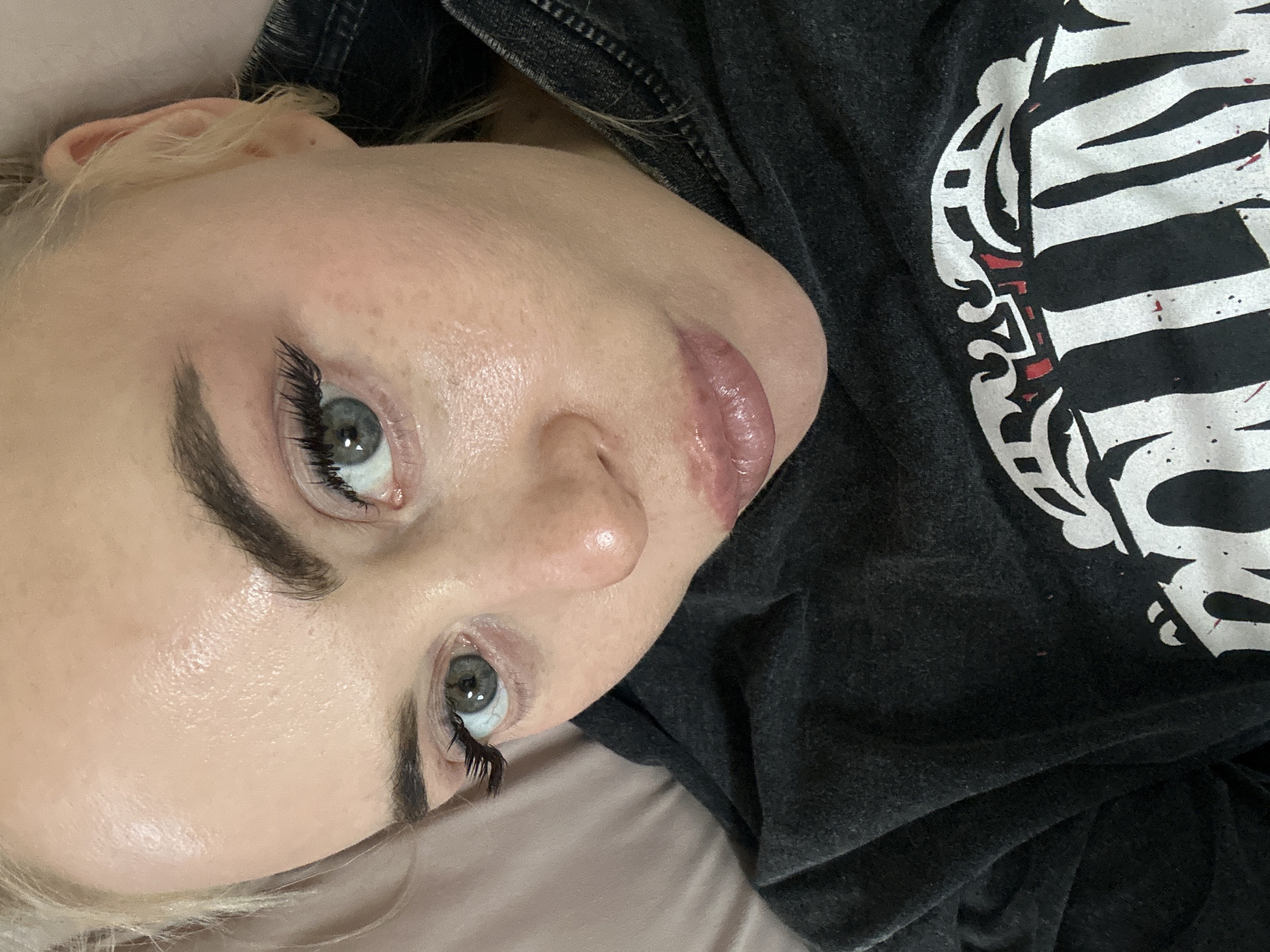 A fair-skinned person with light blonde hair is positioned on a soft surface, likely a bed or pillow, wearing a dark gray or black t-shirt with a white graphic design. The person's face is the focal point, featuring prominent eyebrows, extended eyelashes, and light-colored eyes. The skin appears smooth and possibly slightly dewy, with a natural lip color. The image does not reveal any information about the city of Высокий, Russia. The scene appears to be indoors, well-lit, and the focus is primarily on the person's facial features.