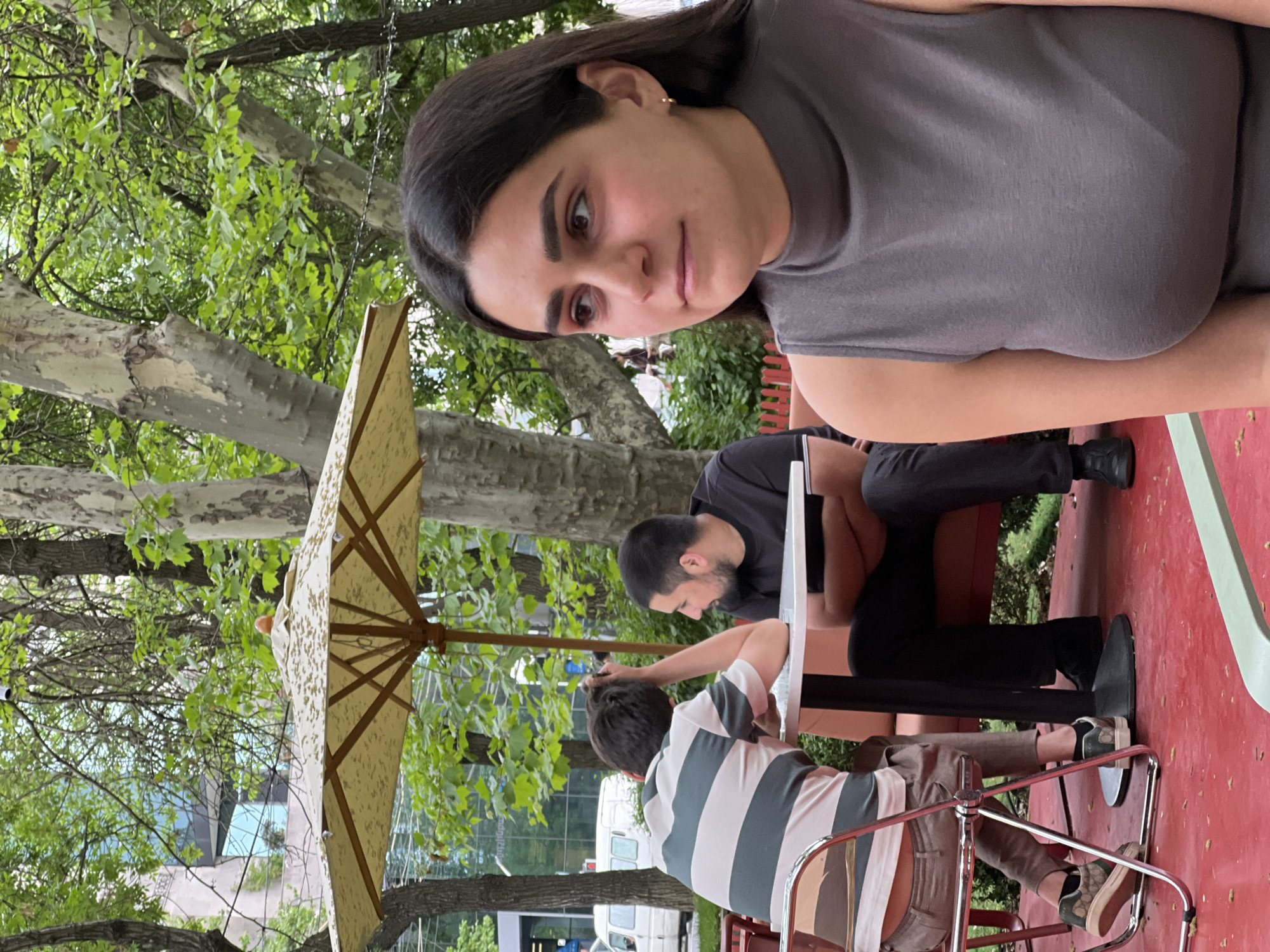 The scene depicts three individuals at an outdoor café in Yerevan, Armenia. A woman with dark hair is prominently featured in the foreground, wearing a grey sleeveless top. Behind her, two men are seated at a table shaded by a patterned umbrella. One man is wearing a black shirt and the other is wearing a striped shirt. The table is on a red surface, possibly a patio or deck. The background consists of trees and a building.
