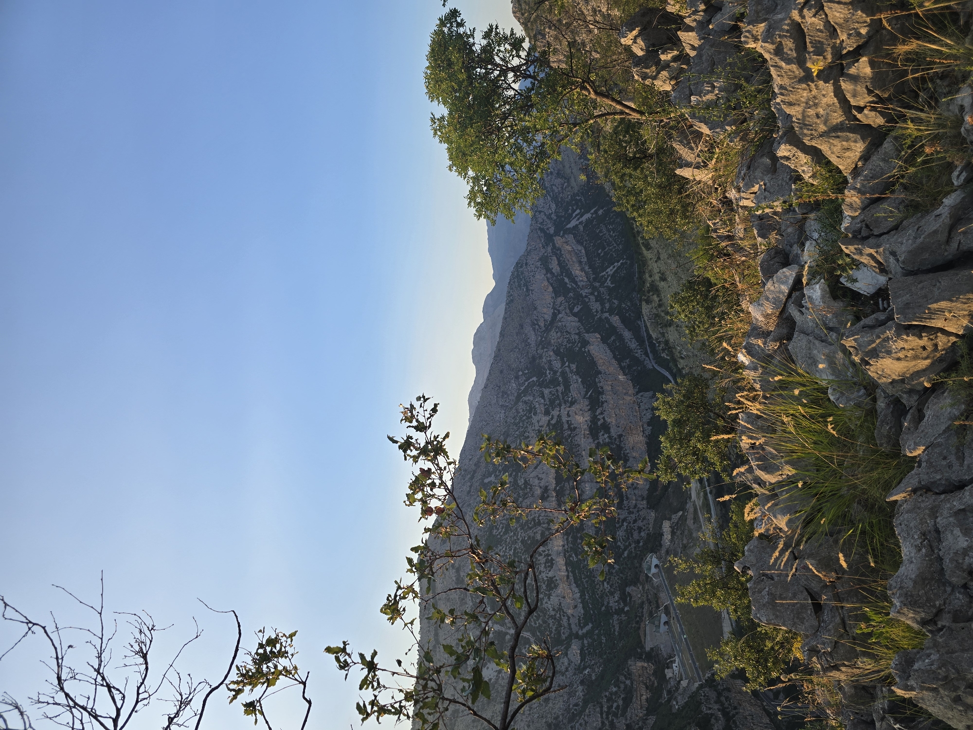 The image captures a scenic view of a mountainous canyon bathed in the warm light of a clear sky, likely during sunrise or sunset. The foreground is dominated by rough, grey rocks and dry grasses, with a few green leaves and branches from trees hanging into the frame on the left. In the mid-ground, a valley with some structures and a road is visible, nestled between steep, rocky slopes. Further in the background, layers of mountains stretch towards the horizon under a pale blue sky. The scene evokes a sense of natural beauty and rugged terrain. There are no people or animals visible in the image. While the image depicts a mountainous landscape, it is not possible to pinpoint a specific location within the city of Podgorica, Montenegro, solely from this photograph without additional contextual information.