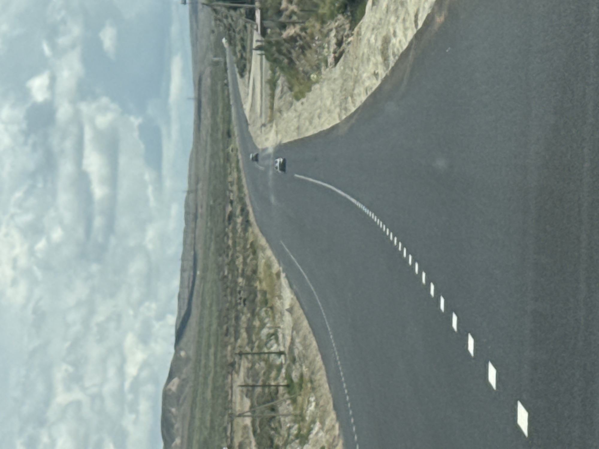 The image depicts a two-lane asphalt road winding through a dry, mountainous landscape. The road is marked with white dashed lines on the right lane and solid white lines on the left. In the distance, two cars are visible on the road. The surrounding terrain is characterized by sparse vegetation, rocky slopes, and distant hills under a cloudy sky. The scene appears to be a rural or remote area, possibly along a highway. There are no identifiable buildings or significant human activity beyond the vehicles on the road. The overall impression is one of travel through a vast, undeveloped natural environment.