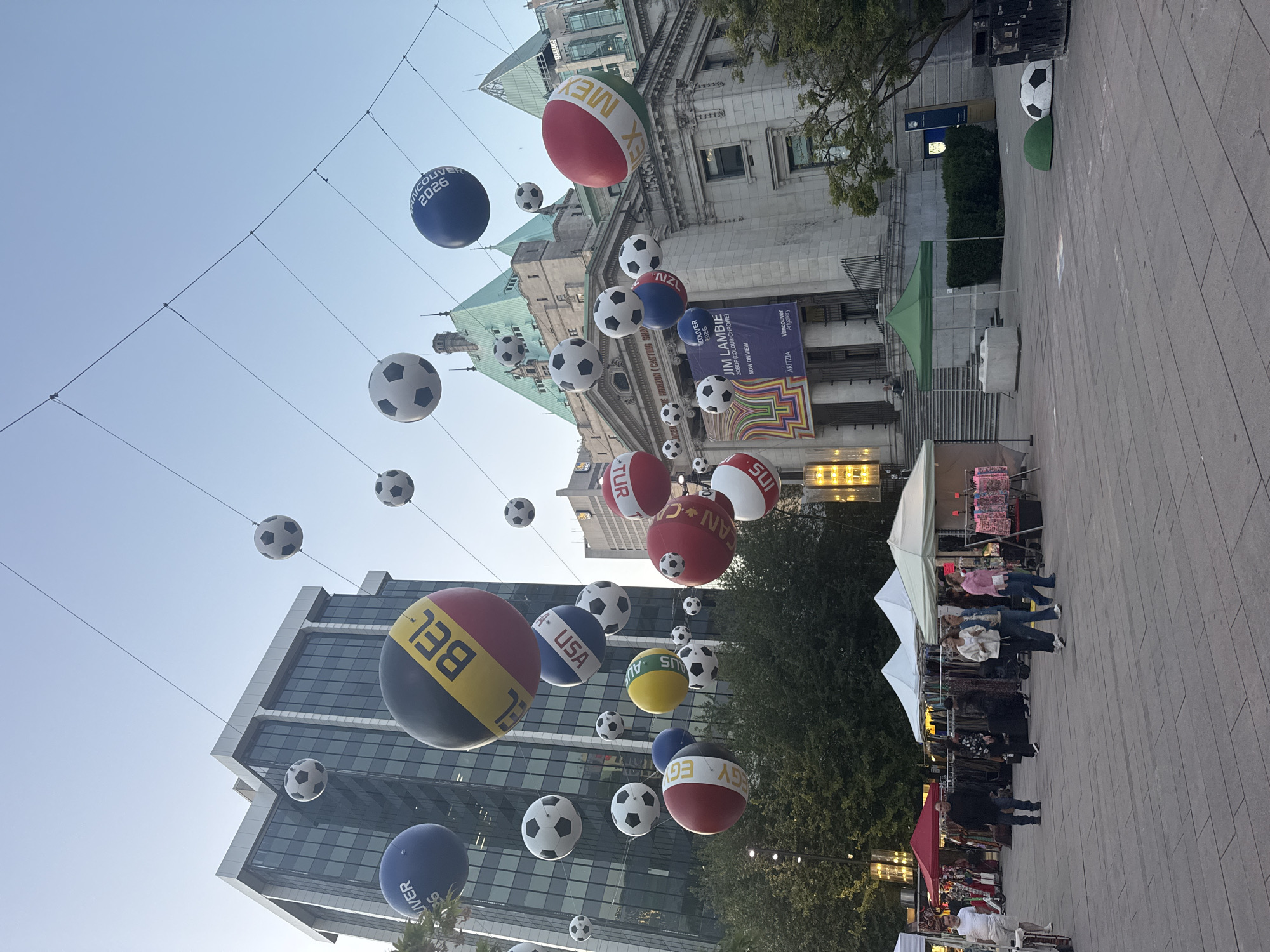 The image shows a street scene in Vancouver, Canada, decorated with soccer balls and country name balloons suspended on wires overhead. People are walking through a market or festival-like setup with tents and stalls on the pavement. A building, likely the Vancouver Art Gallery, is visible in the background, along with a modern glass building. The text "VANCOUVER 2026" is visible on one of the balloons, referencing the city being a host for the FIFA World Cup. A banner for Jim Lambie's "ZOBOP (COLOUR-CHROME)" exhibition at the Vancouver Art Gallery can be seen on the building facade.