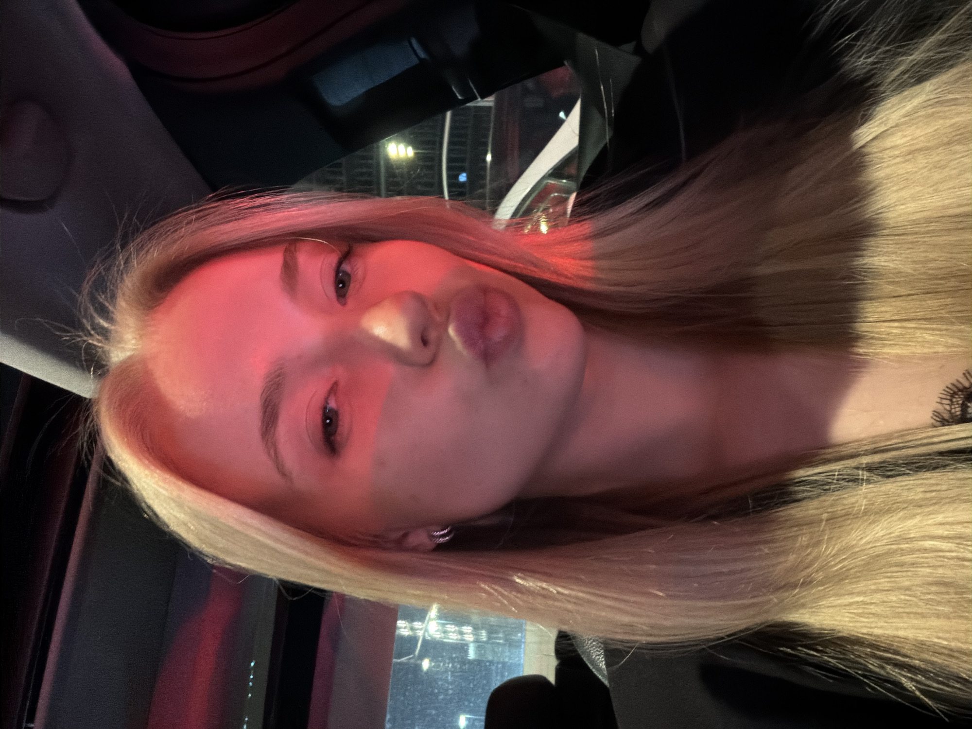 A woman with light blonde hair is posing with pursed lips inside a car. The interior of the car is visible with a dashboard and window. The window shows blurred lights of a city at night. The lighting in the car appears to be reddish.