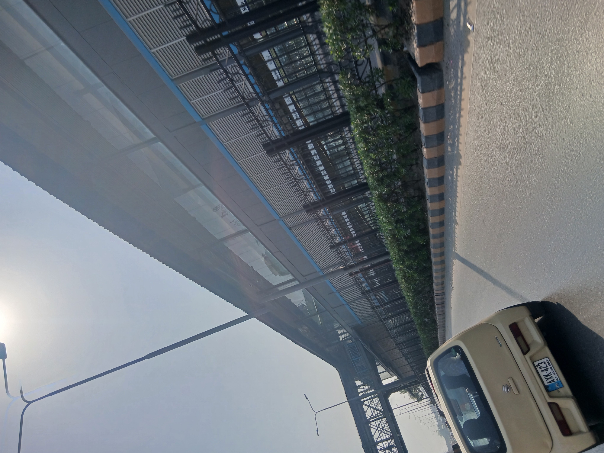 A light yellow, four-door hatchback with a license plate that reads "AXK 423 ICT-Islamabad" is driving on a multi-lane road. The car is angled to the right, showing its rear passenger side. In the background, a modern, elevated structure with a corrugated metal roof and a blue accent line runs parallel to the road. This structure appears to be a transit station or overhead walkway. A pedestrian bridge connects to this elevated structure from the left. The area is lined with manicured green bushes and a curb marked with alternating black and yellow stripes. The sky is bright and hazy, suggesting a sunny day. The scene is set in Peshawar City Tehsil, Pakistan.