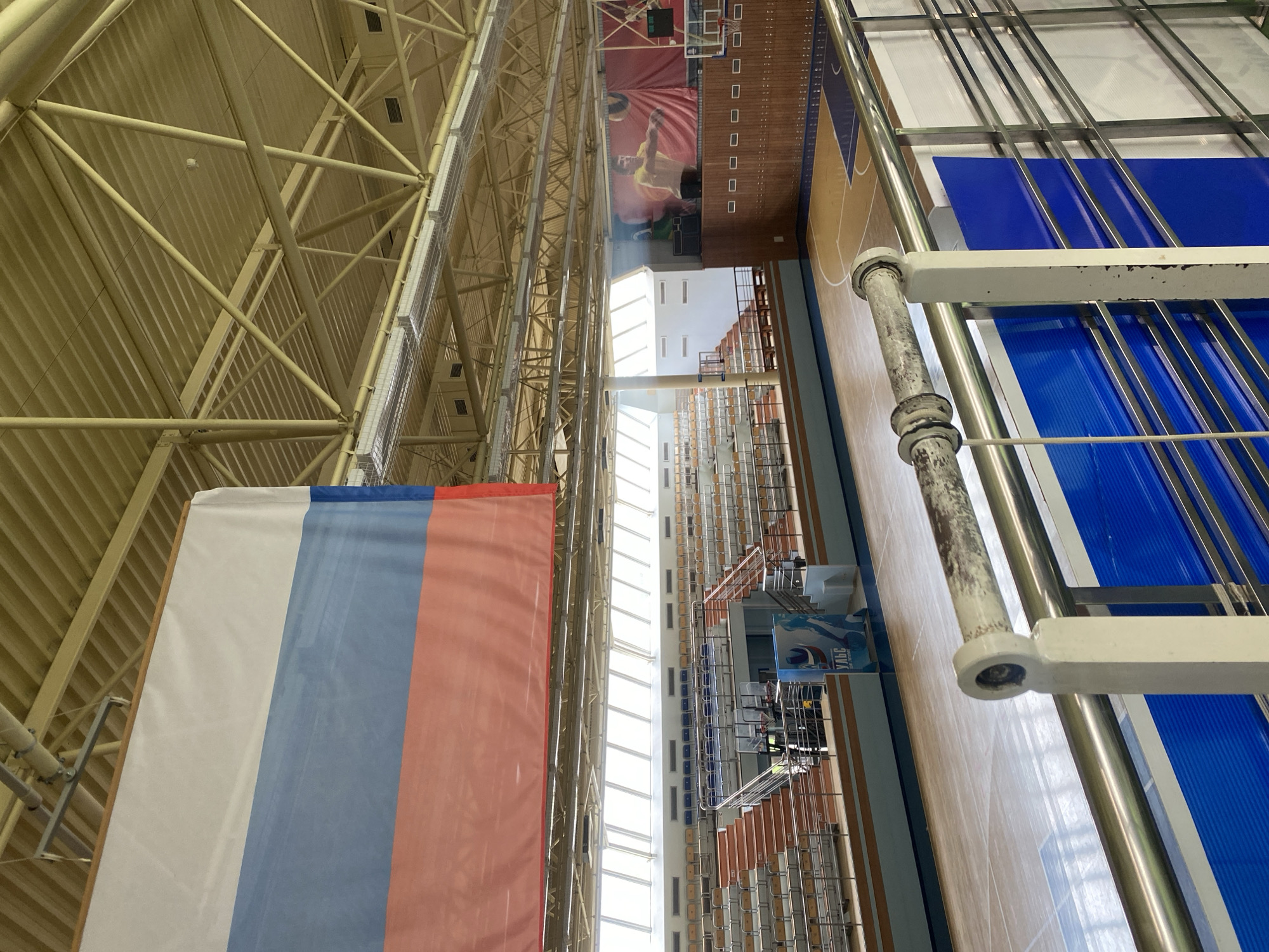 This image captures the interior of a gymnasium in Chaikovsky, Russia. The view is oriented upward, showcasing the building's structural framework, including a complex network of yellow steel beams supporting the roof. Daylight streams in through a long skylight running the length of the building. A large Russian flag hangs prominently from the structure. The gymnasium floor is visible, marked for sports such as basketball, and features bleachers along the side. A banner with an image of a basketball player is displayed high on the end of the gymnasium. No people are distinctly visible in the image, though there is a possibility of people being present along the wall in the lower right.