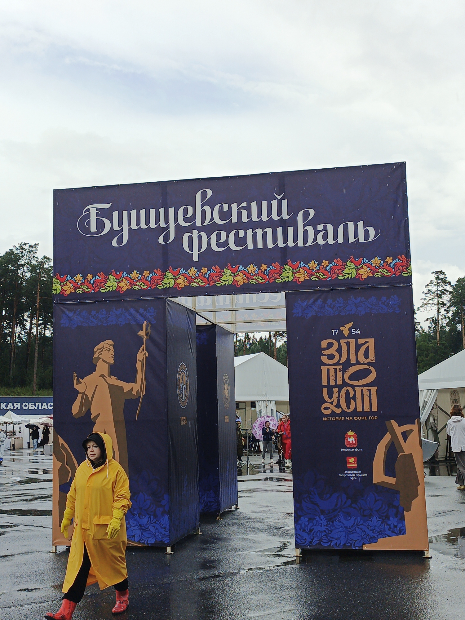 The image depicts an outdoor scene with a large archway adorned with a dark blue banner featuring Cyrillic text. The banner's text reads "Бушуевский фестиваль," and it is decorated with colorful floral motifs. Beneath the main banner, on the sides of the archway, there are dark blue panels with blue floral patterns. On the left panel, a stylized golden figure of a person holding a staff is visible. The right panel displays a vertical graphic with the text "17 54 Златоуст ИСТОРИЯ НА ФОНЕ ГОР," indicating the city of Zlatoust and its history. Below this text are two official emblems, likely of the Chelyabinsk Oblast and Zlatoust city.
In the foreground, a person wearing a bright yellow hooded raincoat, yellow gloves, and red boots is walking from left to right across a wet, paved surface, suggesting it is raining or has recently rained. The ground reflects the light. In the background, several white tents and trees are visible, indicating an event or festival setting. Other people are partially visible in the background, some holding umbrellas. A banner with the text "КАЯ ОБЛАСТ" is also visible on the left side of the frame. The overall atmosphere suggests an event taking place in Zlatoust, Russia, possibly during inclement weather.