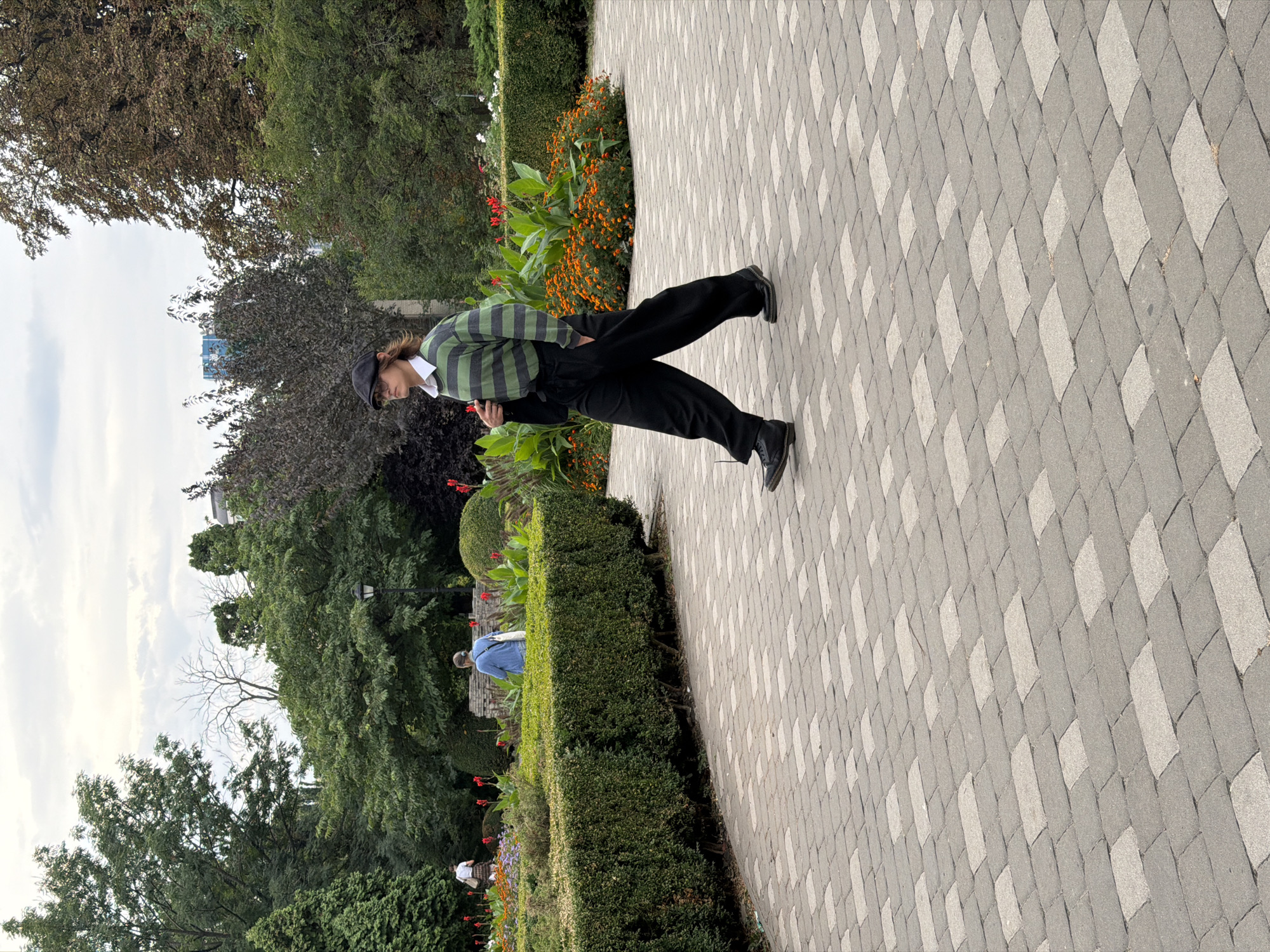 The image shows a person walking on a paved path in a park. The person is wearing a green and black striped sweater, black pants, a black cap, and black shoes. They are carrying a black jacket over their shoulder. In the background, there are manicured bushes, flower beds with orange and red flowers, and trees. Two other people are visible in the background, one in a blue jacket and the other in a white top. The sky is overcast with clouds. The location is the Mariinskyi Park in Kyiv, Ukraine. The park features a formal garden with hedges and colorful flowers, and the paved walkway is made of rectangular stones. The overall scene depicts a calm day in a city park, with a focus on the lone individual walking through the grounds.