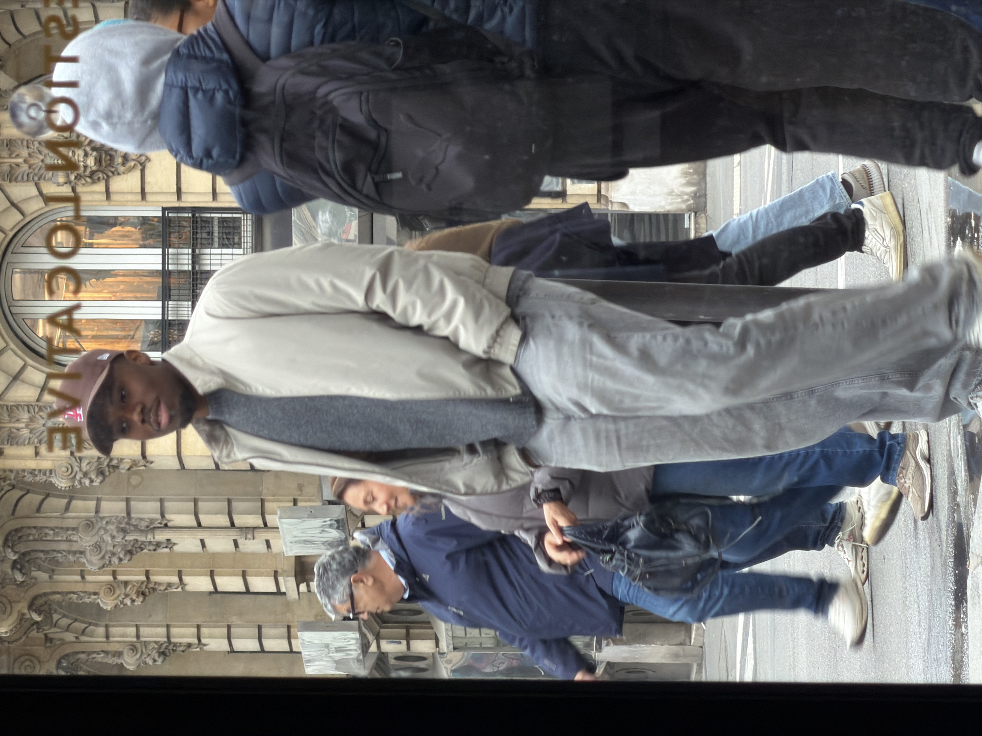 The image is a street scene in Paris, France, reflected in a window. Several pedestrians are visible: a man in a tan jacket and cap, a woman in a grey jacket, and a man in a blue jacket. The architecture in the background is detailed with decorative carvings and windows. The reflection suggests a typical urban environment.