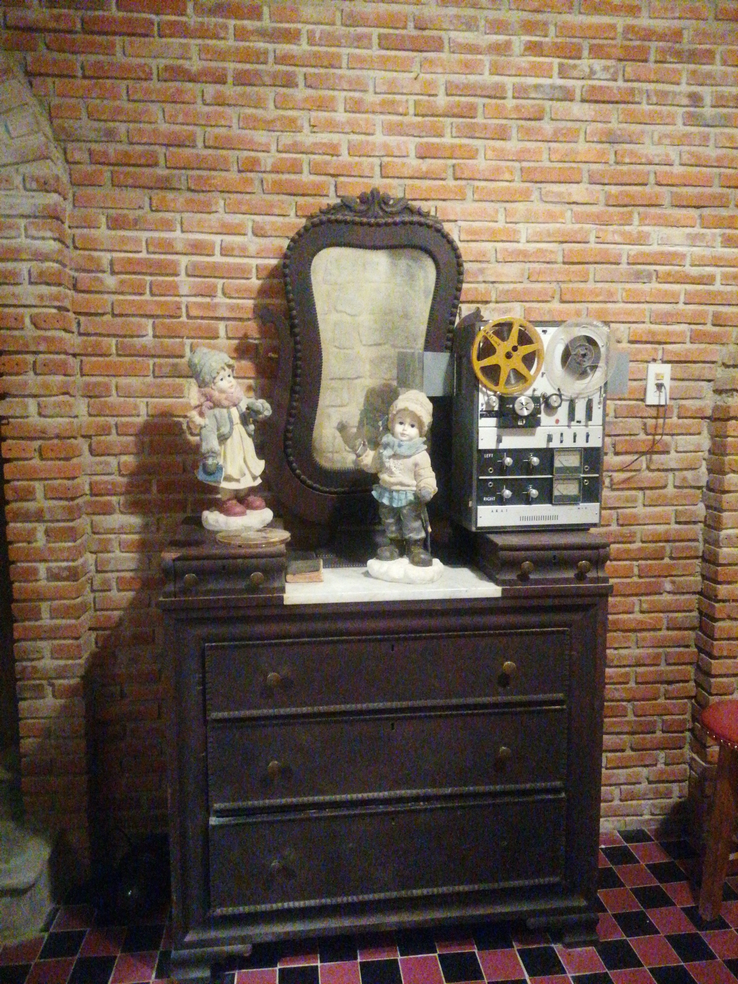 The image depicts an indoor scene with a brick wall as the backdrop. In the foreground, a dark wood dresser stands against the wall. On top of the dresser, there are several objects. To the left, there is a figurine of a child wearing a hat and coat, standing on what appears to be a cloud-like base. Next to it, to the right, is a reel-to-reel tape recorder with a yellow reel and a transparent reel. Behind these objects, a mirror with an ornate, dark frame is positioned against the brick wall. The floor is tiled with a pattern of dark and reddish squares. The overall impression is of a vintage or retro display, potentially in a shop or a home. No specific location in Da Lat, Vietnam, can be identified from the image itself.
