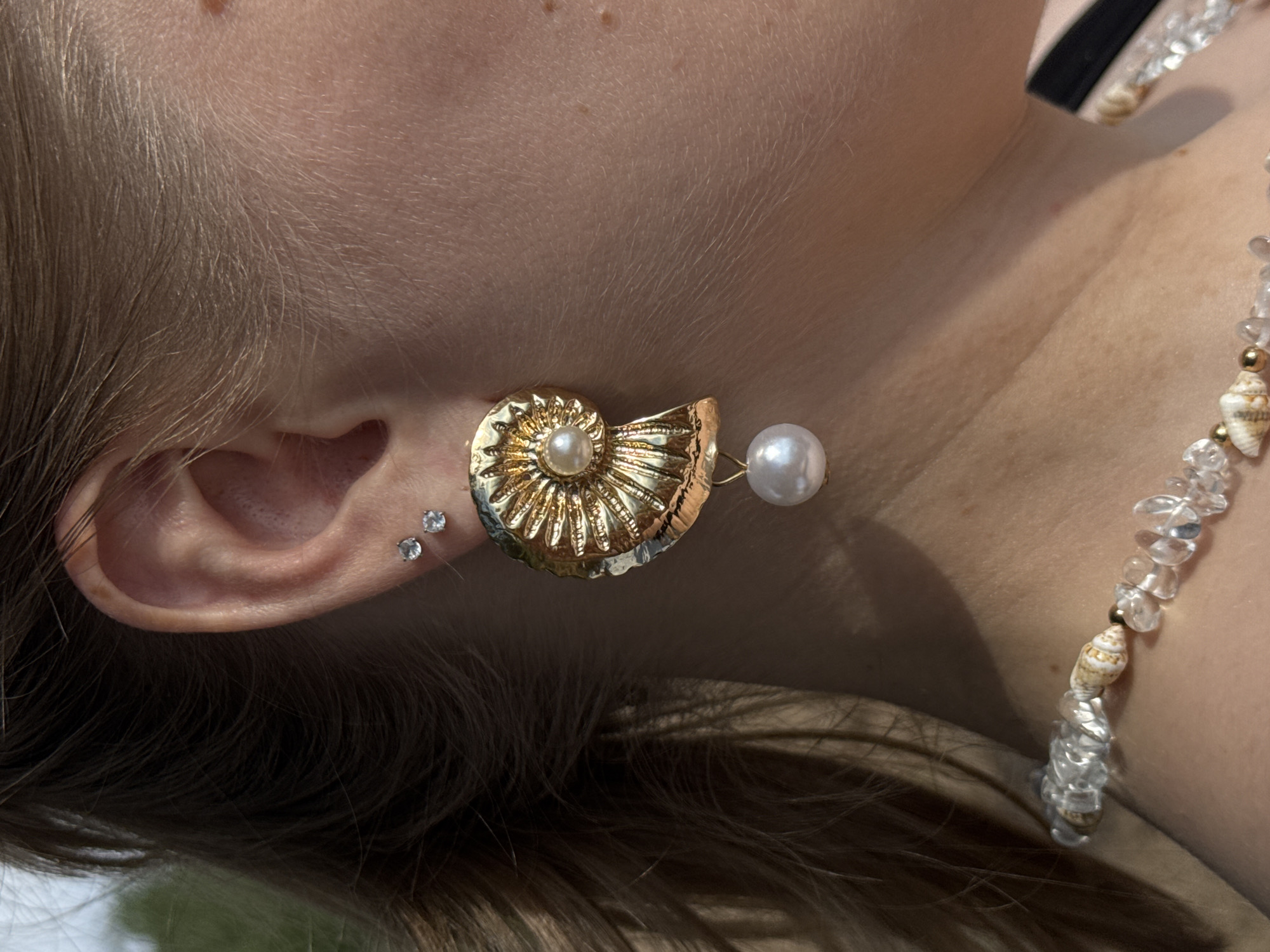 This media file features a close-up shot of a person's ear, neck, and shoulder, adorned with jewelry. The primary subject is the ear, with a prominent gold, shell-shaped earring featuring a pearl. Next to the earring, two small, sparkling studs are visible on the earlobe. The person's neck and shoulder are also visible, showing a necklace made of clear crystals and small seashells. The lighting suggests it is daytime, with natural light illuminating the scene. The background is softly blurred, showing hints of greenery, which could indicate an outdoor setting. The focus is on the intricate details of the jewelry and the textures of the skin and hair. No text is visible in the image.
