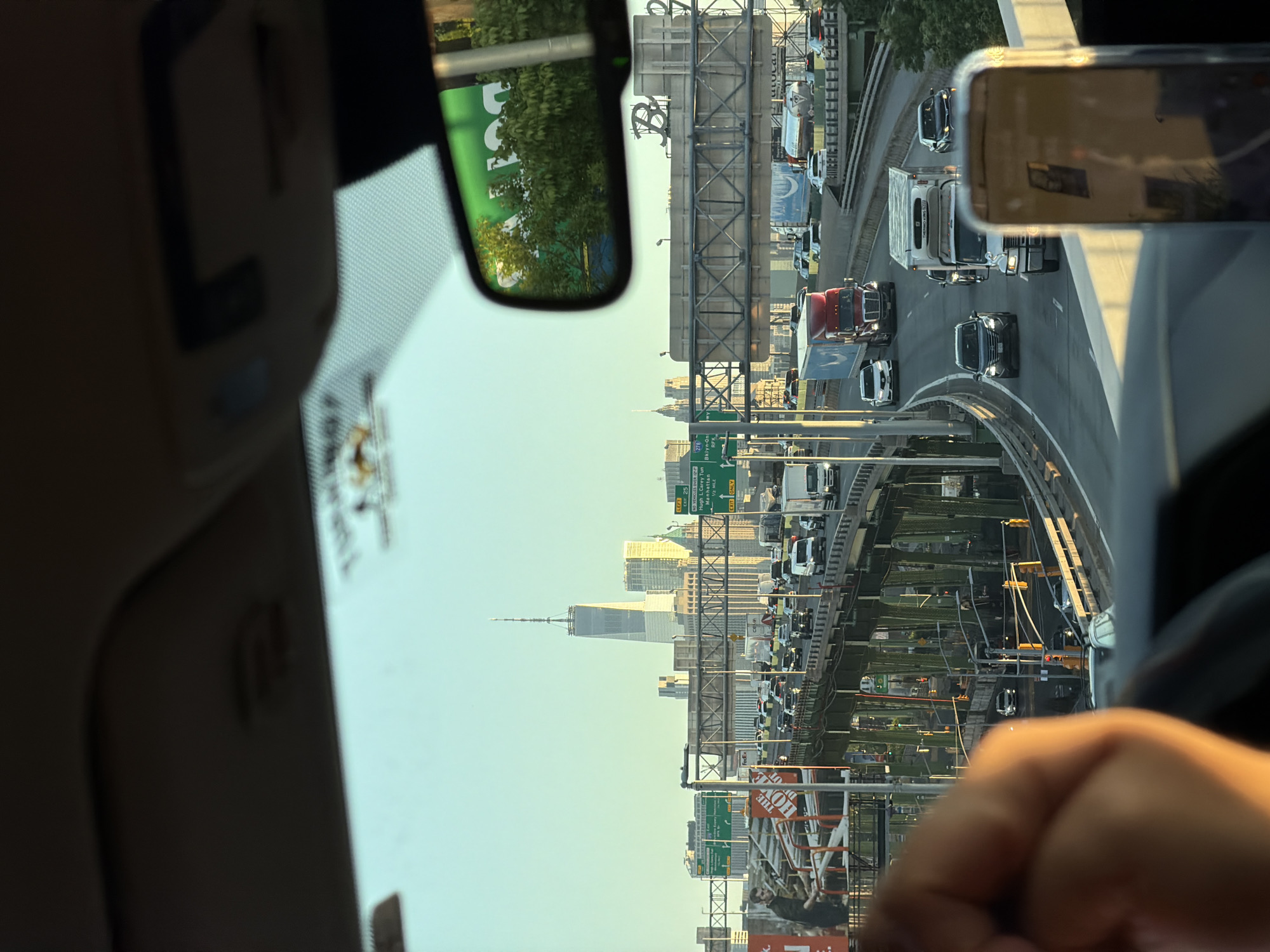 The image, taken from inside a vehicle, shows a view of traffic on a highway in New York City, United States of America. The One World Trade Center skyscraper is visible in the distance. A rearview mirror and a mobile phone are visible within the vehicle's interior. Traffic is dense. Road signs indicate highway exits.