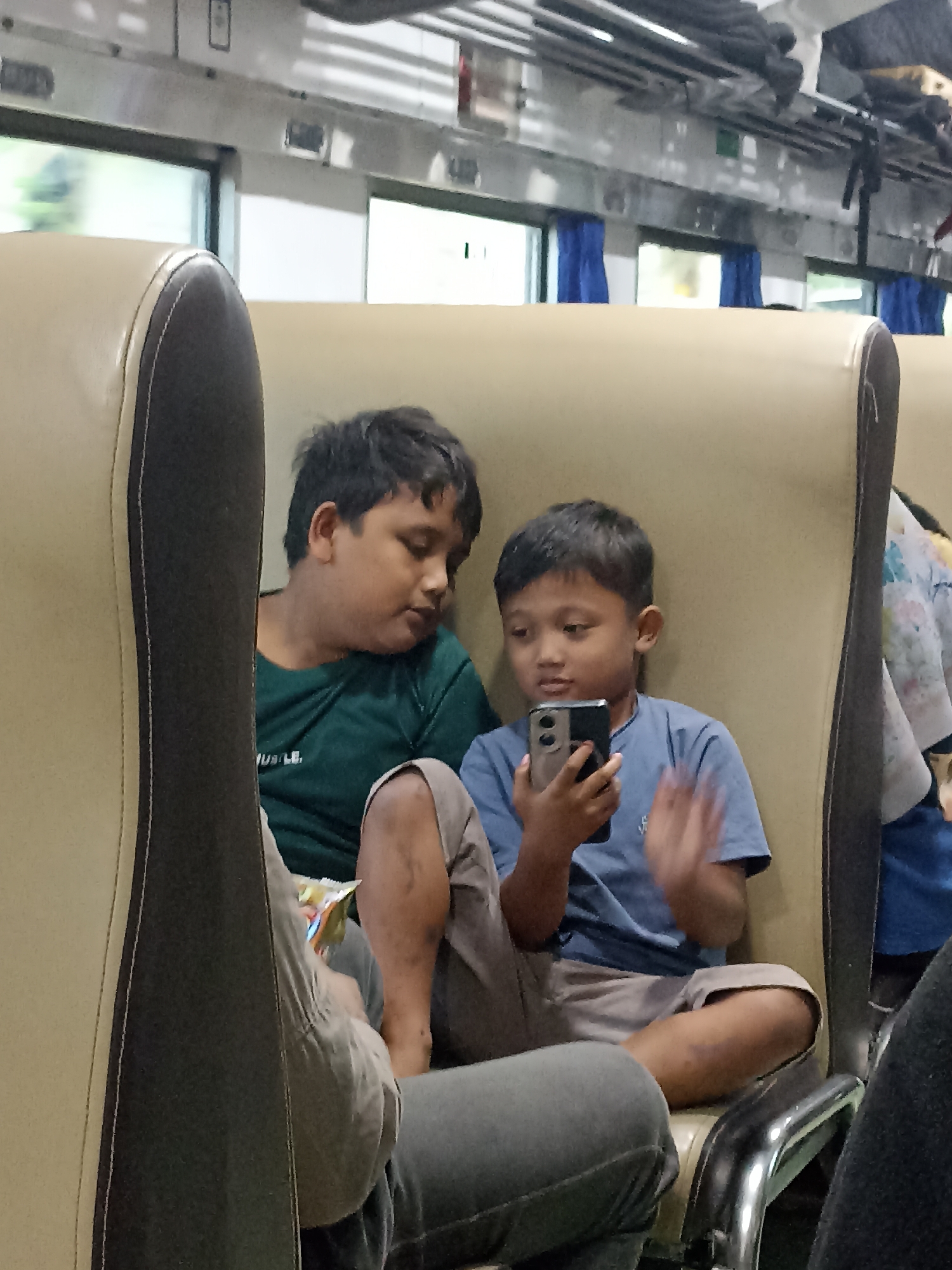 Two young boys are seated next to each other on a train. The boy on the left, wearing a green t-shirt, is looking down. The boy on the right, wearing a blue t-shirt, is holding a smartphone and appears to be interacting with it, possibly playing a game or watching a video. He is looking intently at the screen. The train interior is visible with beige and dark brown seats, windows with blue curtains, and overhead storage. The scene is illuminated by ambient light, possibly from the windows and interior lights. The location is identified as being in the city of Jambewangi, Indonesia.