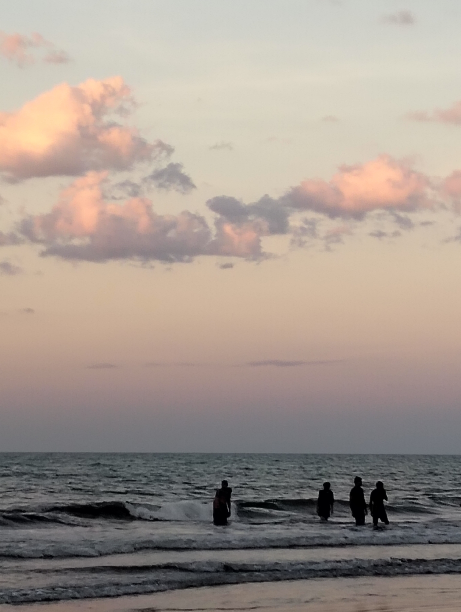 This is a vertical, eye-level shot of a beach scene at dusk in Peukan Sot, Indonesia. The sky is a gradient of pale blue to soft pink and peach, with several wispy clouds tinged with pink and gray. The ocean occupies the lower half of the frame, with gentle waves breaking onto the shore. Several silhouetted figures, appearing to be people, are standing in the shallow water. They are facing the ocean, with some appearing to be interacting with the water. The overall mood is serene and peaceful, typical of a sunset at the beach.