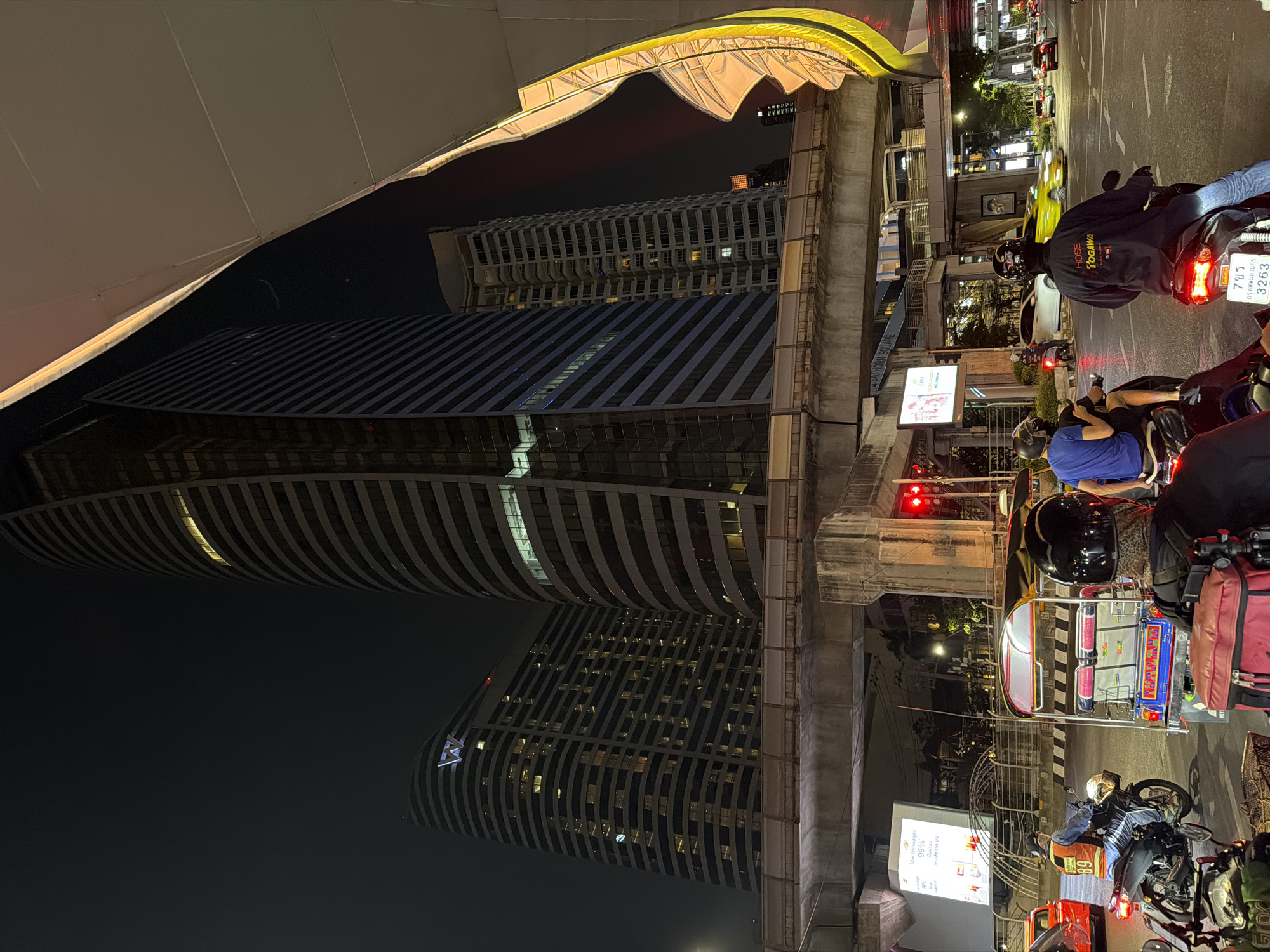 The image captures a nighttime street scene in Bangkok, Thailand. A tall, modern building with a unique curved design dominates the left side of the frame. The architecture suggests a contemporary hotel or office structure. The scene is illuminated by a combination of building lights and streetlights.
On the right side, the elevated train tracks are visible, supported by concrete pillars. Below the tracks, there is a busy street with several motorcycles and a tuk-tuk waiting at a red light. The motorcycles are a mix of standard bikes and scooters, and the tuk-tuk is brightly decorated, typical of Thailand. Riders are wearing helmets.
Billboards with Thai writing are visible near the street, adding to the local atmosphere. The scene suggests a typical busy intersection in Bangkok at night. The presence of both modern architecture and traditional vehicles highlights the blend of old and new in the city.
