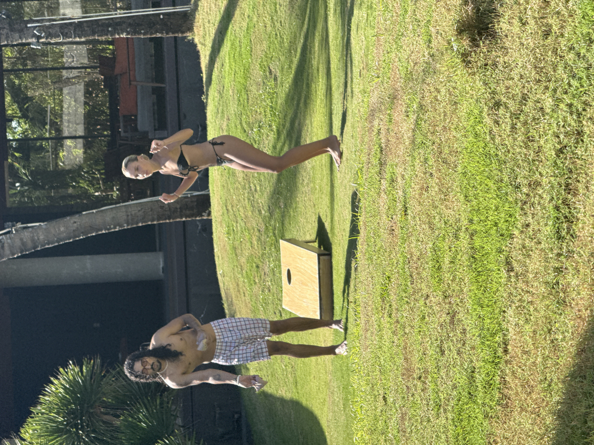 Outdoors in Đà Nẵng, Vietnam, a man and a woman stand on a grassy lawn. The man, shirtless and wearing plaid shorts, throws a beanbag. The woman, wearing a black bikini, throws a beanbag as well. A cornhole game is set up on the lawn between them. Palm trees and building facades are visible in the background.