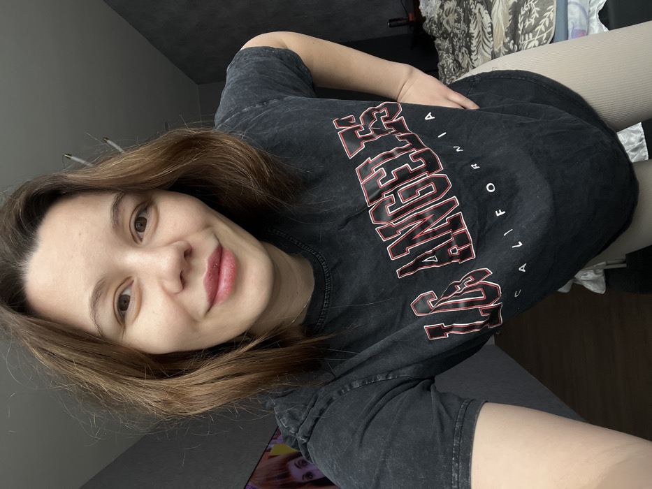 The image depicts a young adult female positioned centrally, facing forward and slightly to the viewer's right. She has brown hair, fair skin, and a slight smile, with her eyes open and directed at the camera. She wears a faded, dark grey or black short-sleeved t-shirt featuring "LOS ANGELES CALIFORNIA" printed in red and white block lettering across the chest. Her right hand rests on her hip, and the lower portion of light beige, ribbed leggings is visible on the bottom right. The background on the left consists of a dark grey, textured wall, with a partial view of a television screen displaying an animated character with orange hair and prominent eyes. The ceiling is light-colored. On the right, a section of a bed with a dark-patterned duvet or blanket and a dark, slender object on a surface next to the bed are visible. The specific location in the city of Leninakan, Russia, cannot be determined from the visual information provided in the image; Leninakan is historically located in Armenia.