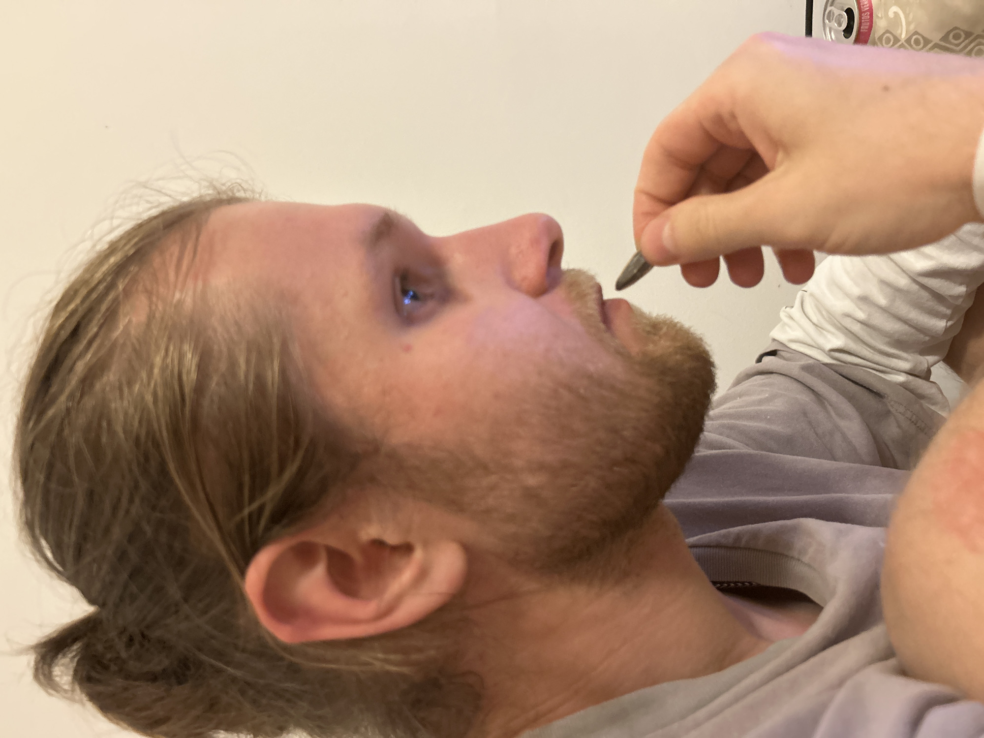 A close-up shot depicts a light-skinned person with blond hair pulled back in a low bun and a reddish-brown beard. The person is in profile, looking upwards. A hand is holding a dark-colored object near the person's mouth. The person is wearing a gray t-shirt with a white long-sleeved shirt underneath. The location is not discernible from the image.