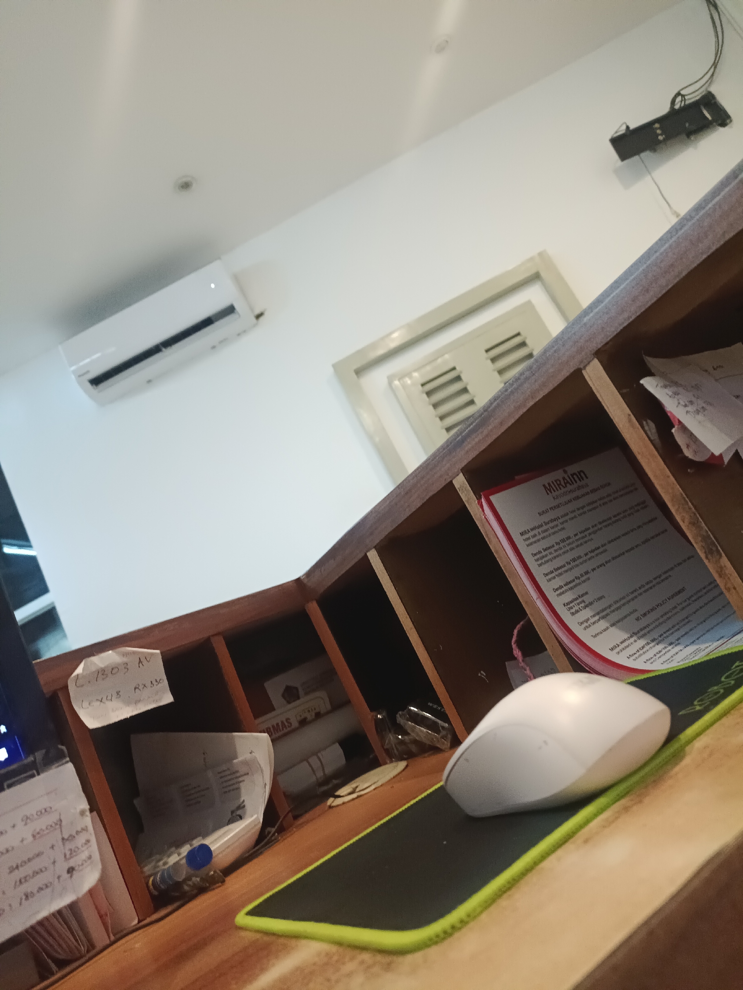 Here is a description of the image:
The image shows a cluttered desk or counter space. On the desk is a white computer mouse on a black mousepad with a bright green trim. Behind the mouse are various papers and documents, some in a wooden cubby shelf. One document visible on the counter has "MIRAINN" printed on it in large letters and has smaller print underneath. Another paper in the cubby reads "L.1303 AV" and "LEXUS RX330". Other papers and small bottles are also on the desk. On the wall is an air conditioning unit. The area appears to be indoor.