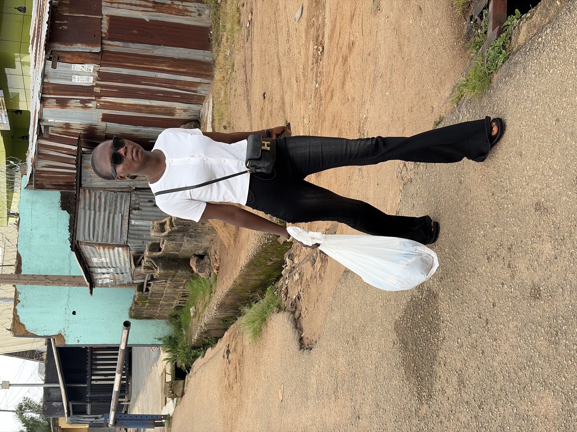 A full-body, eye-level shot captures a Black woman walking on a dirt road in Jos, Nigeria. She is wearing a white, short-sleeved, collared shirt with buttons, black flared jeans, and black sandals. A small, black, crocodile-textured handbag with a gold "H" clasp hangs from a black strap across her body. She is holding a large, translucent white plastic bag filled with items in her right hand. Sunglasses with dark lenses are perched on her nose. The background features a rustic, corrugated metal structure and a turquoise-colored building. A narrow concrete ditch runs along the right side of the road, bordered by overgrown grass and some exposed concrete blocks. The road surface is rough and uneven, with patches of dirt and gravel. The overall scene depicts an everyday moment in a somewhat underdeveloped urban or semi-urban environment.