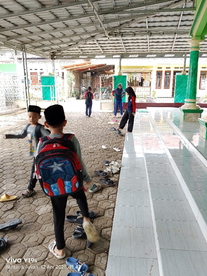 The image shows a group of children in Anjatan, Indonesia, located at coordinates -6.419 latitude and 107.979 longitude. The children are all wearing school uniforms and are standing in front of a building. The building appears to be a mosque. The image is taken from the perspective of a person standing on the steps leading up to the building. There are a few sets of shoes left on the steps. Some children are wearing backpacks. There is a green column behind the children. The area appears to be an outdoor courtyard with a concrete floor and a metal roof. The children are interacting with each other and appear to be chatting. The environment is relatively quiet, and the children are in a relaxed and friendly atmosphere.