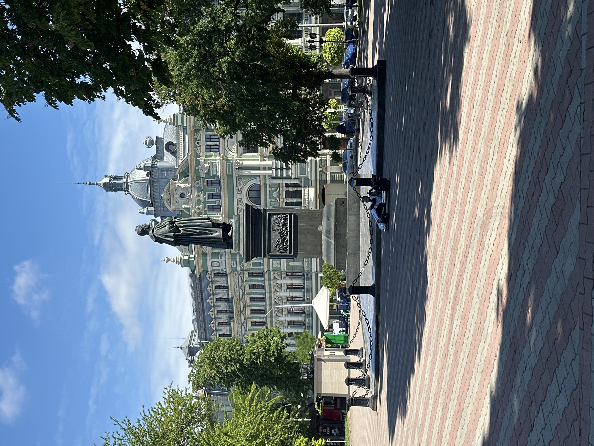 Here is a detailed description of the image:
The image shows a statue in front of a building with ornate architectural details in an urban environment. The statue, depicting a robed figure on a pedestal, dominates the view and stands prominently against the backdrop of the building. The building itself exhibits a complex facade, featuring a dome, multiple windows, and decorative elements.
The environment includes trees that frame the scene on either side, adding a touch of greenery. The foreground consists of a paved walkway, its pattern of interlocking stones creating a sense of depth. Chains are visible near the statue, possibly serving as a barrier.
Two people are visible in the bottom of the frame, they are sitting on the base of the statue.
The location is in Unknown, Unknown.