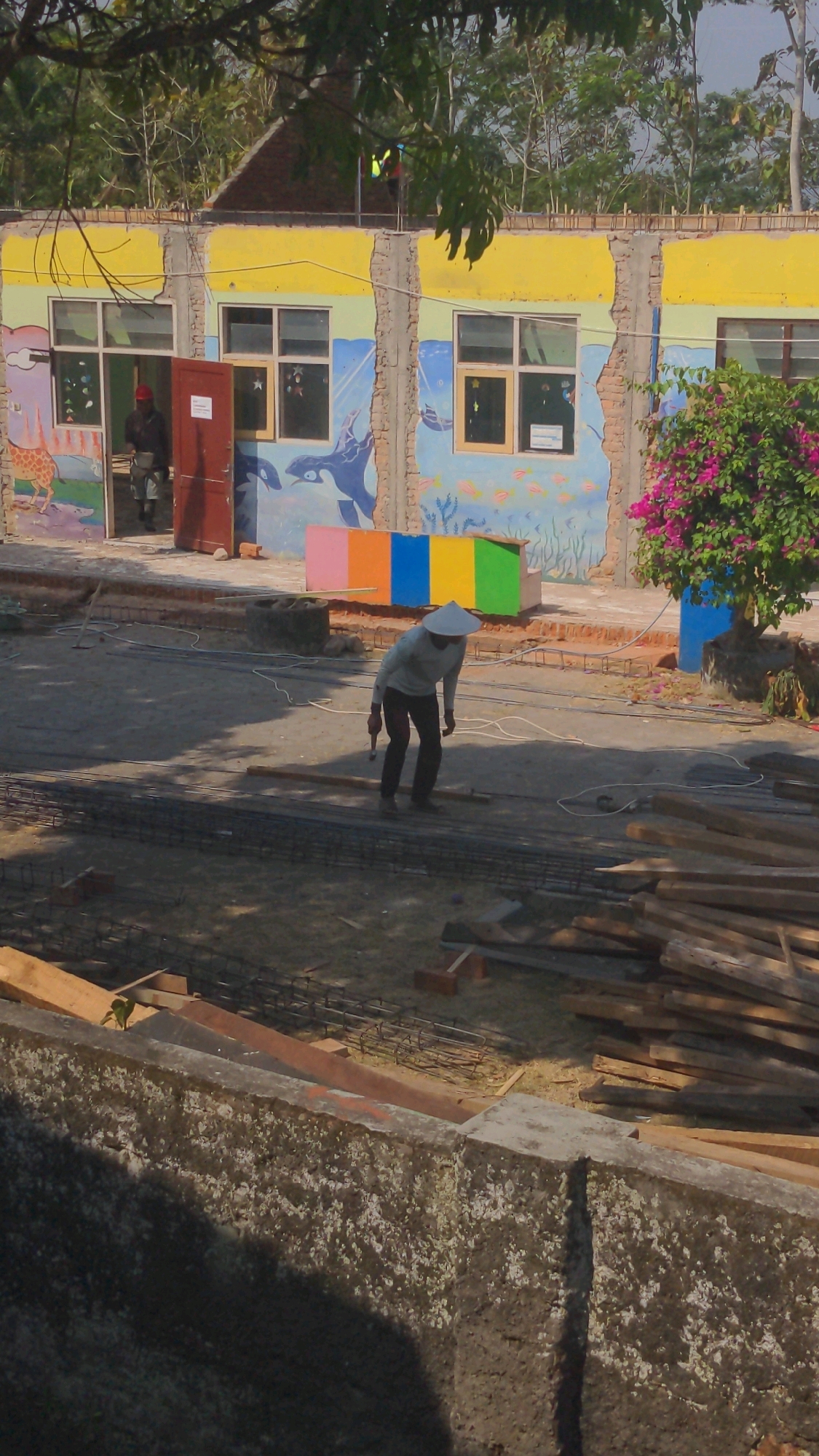 In Tembarak, Indonesia, a construction site is depicted with a partially constructed building in the background. The building features yellow trim and a mural of aquatic animals on its exterior walls. A person wearing a conical hat and dark clothing is working on the ground in front of the building, which is a cement structure. The construction area is littered with building materials, including rebar and wooden planks.
