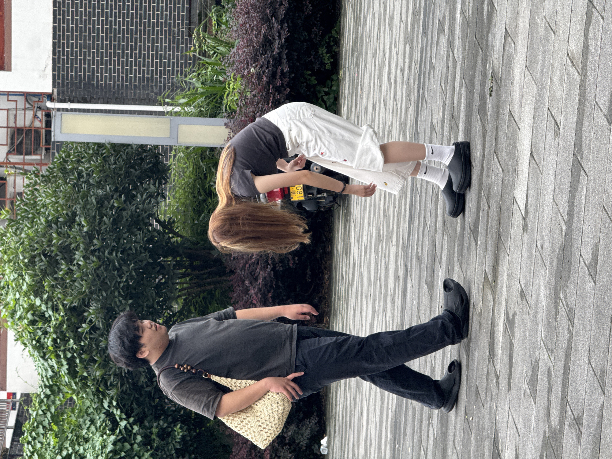 In Shapingba District, China, on a paved walkway, a man and a woman are standing outdoors. The man, on the left, is wearing a dark t-shirt, dark pants, and dark clogs. He is carrying a light-colored woven bag over his left shoulder. The woman, on the right, is wearing a dark t-shirt, white shorts, white socks, and black clogs. She is bending forward, with her head down, as if looking at something on the ground. Behind them, there are green bushes and a brick wall. A motorcycle is partially visible in the background.