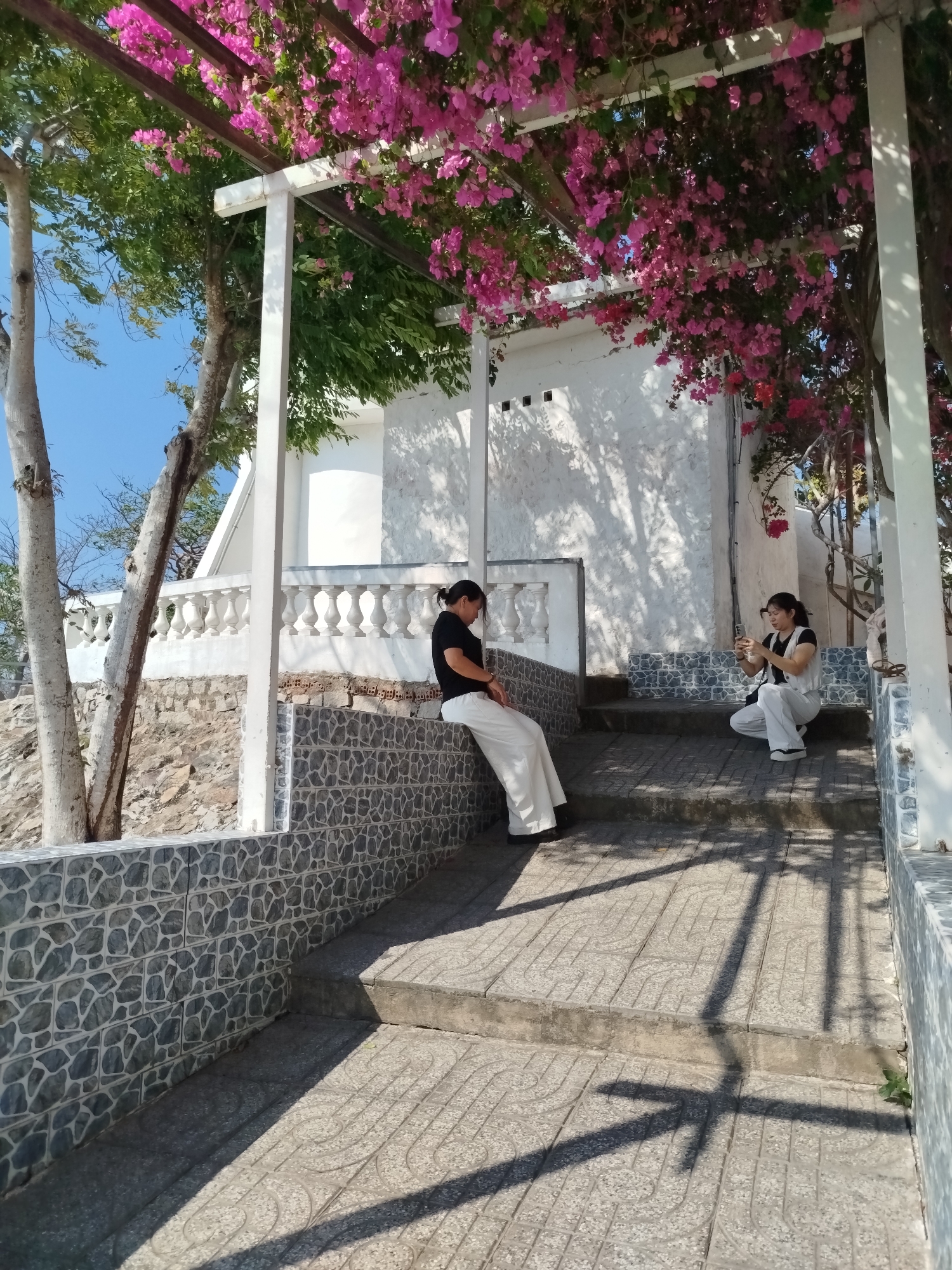 The image shows two women on a tiled stairway leading to a white building under an arbor covered in pink flowers. The woman on the left is seated on a tiled wall and wearing white pants and a black shirt. The woman on the right is crouching on the stairs, wearing white pants, a white vest, and a black shirt, and holding a smartphone. The setting appears to be outdoors on a sunny day in a location with stone walls, tiles, and flowering plants. The location could be in Ho Chi Minh City, Vietnam.