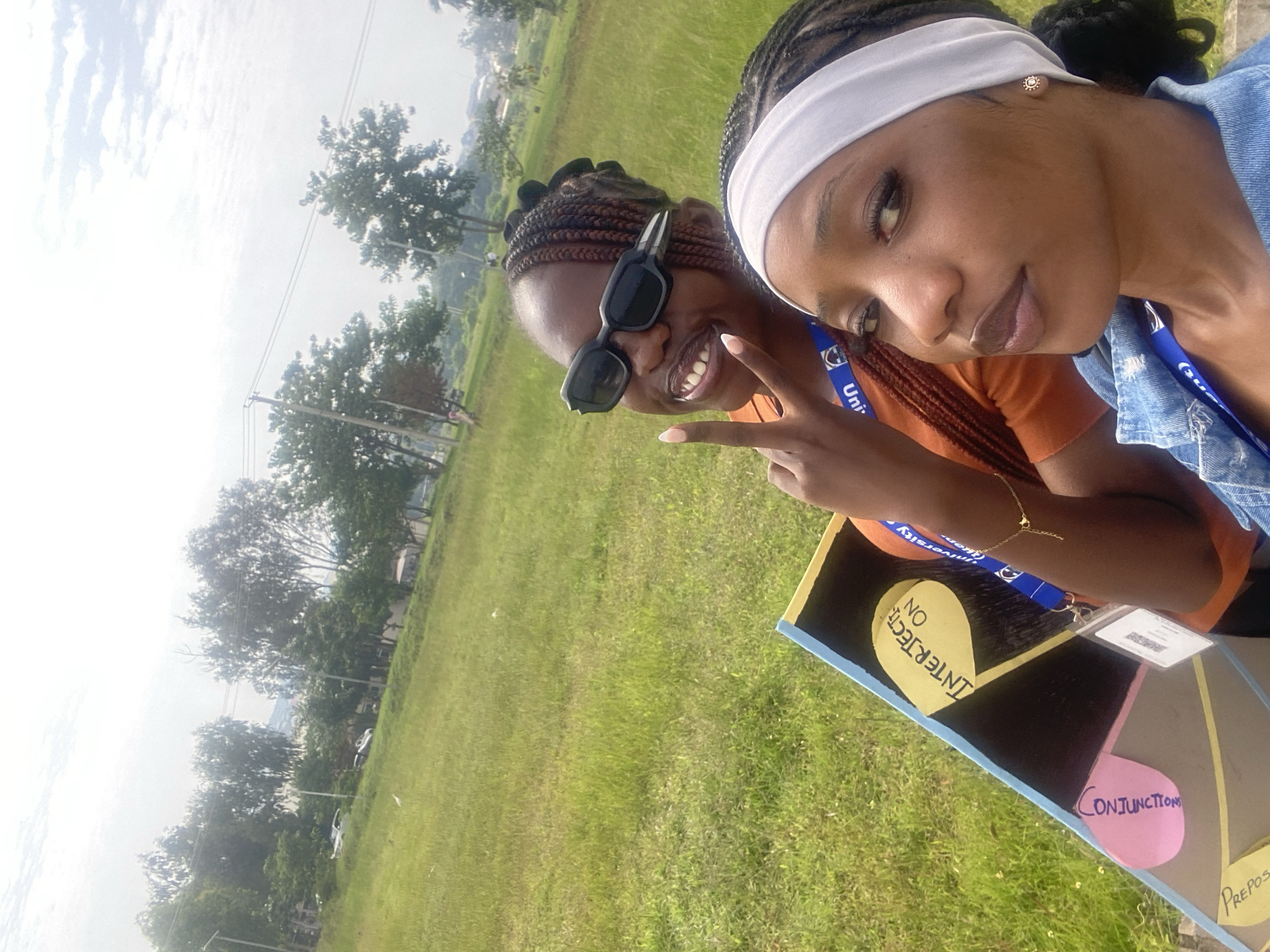 This is a close-up, selfie-style photograph taken outdoors on a bright day. Two young Black women are featured. The woman in the foreground, on the right, is wearing a white headband, a blue denim jacket, and has dark hair braided into a bun. She is looking directly at the camera with a neutral expression. Her left ear is adorned with a small, circular stud earring.
Behind her, on the left, is another young Black woman wearing black, angular sunglasses and an orange top. She is smiling and holding up two fingers in a peace sign. Her dark hair is in braids. A blue lanyard with a white "Uni..." logo is visible around her neck.
Both women appear to be in a grassy area, likely on a university campus given the lanyard. In the background, there are trees, a paved area with a few parked cars, and buildings in the distance. The overall impression is one of a casual, outdoor gathering or event. The photograph is likely taken in Jos, Nigeria, considering the context of similar images from the region. The weather appears to be partly cloudy.