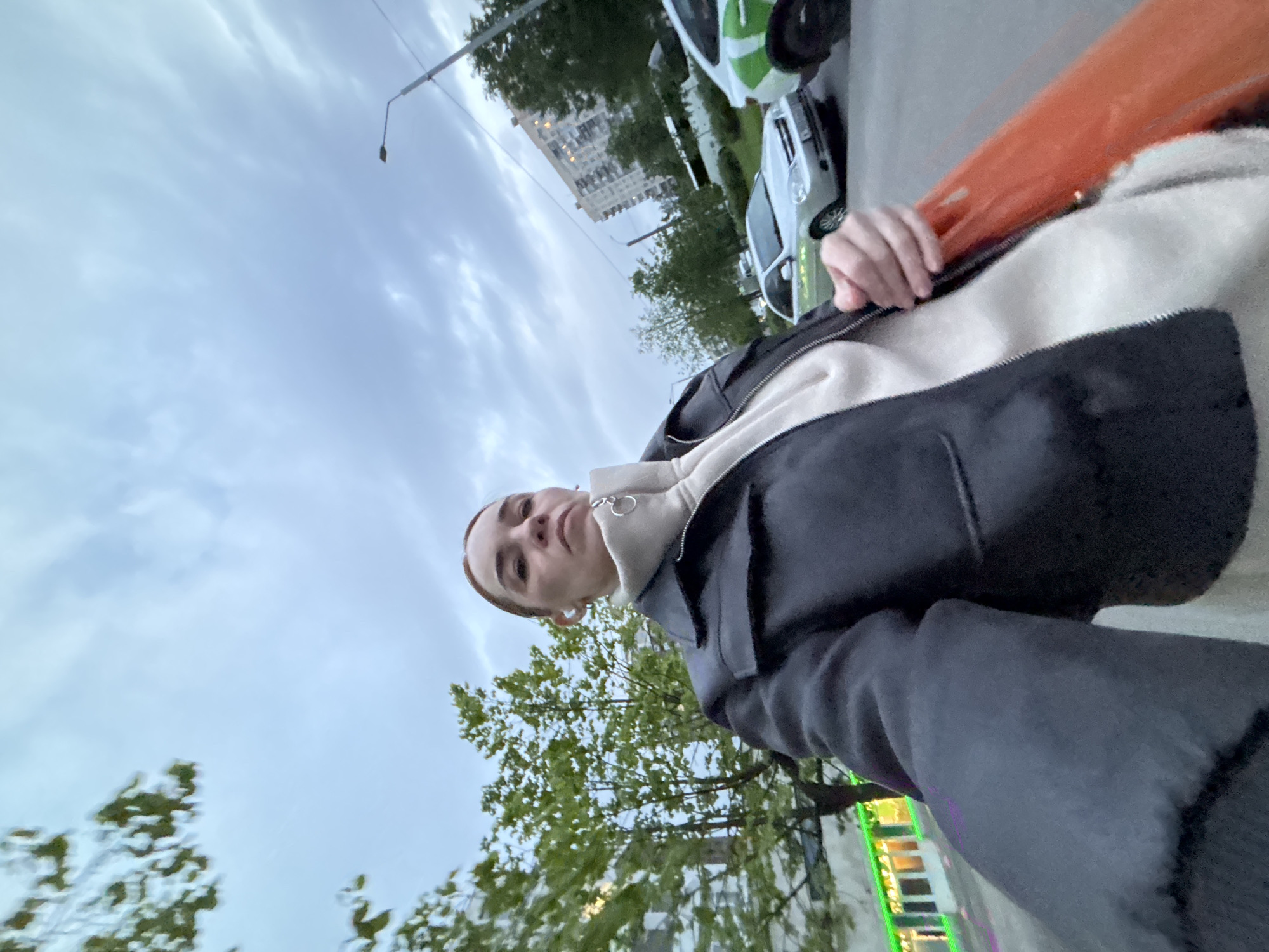 A downward-angled outdoor shot shows a person in a black jacket over a light-colored hoodie in an urban setting. In the background, there are trees, a light pole, and buildings. Two cars, one white and one white with green markings, are partially visible. The sky is overcast. A brightly colored object is partially visible, held by the person. A building with illuminated signs is also in view.