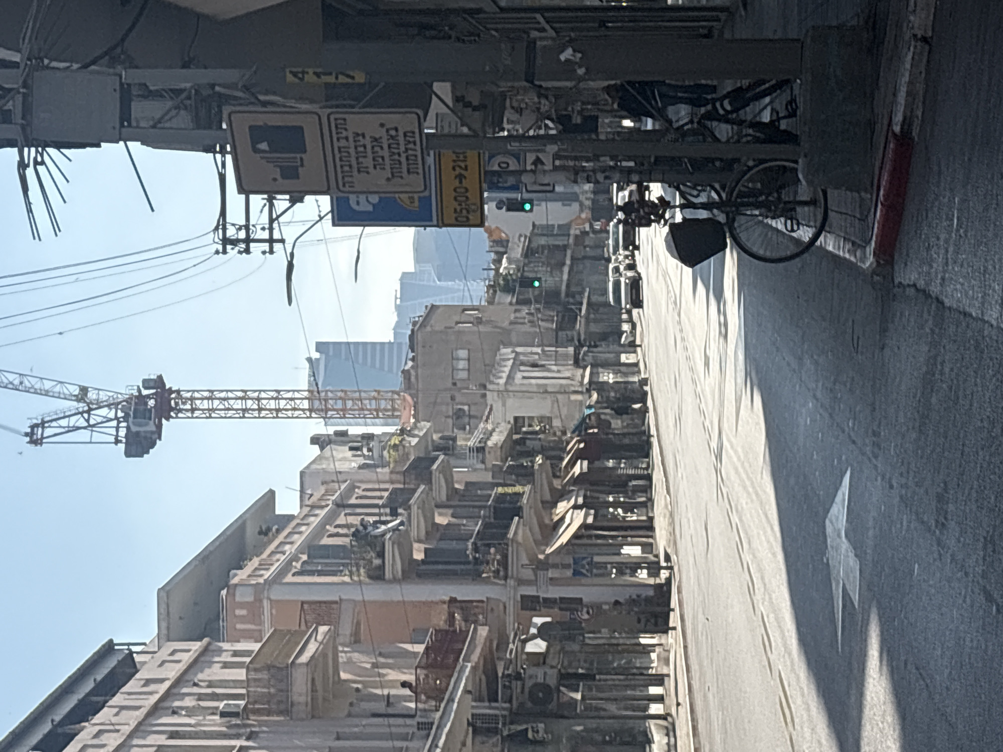 The image shows a street in Tel-Aviv, Israel. The street is narrow and lined with buildings of various heights, many with visible air conditioning units. A construction crane is visible in the background. Street signs are in Hebrew, with one indicating time restrictions from 05:00 to 21:00. A bicycle is parked on the right side of the street. An arrow is painted on the street surface.