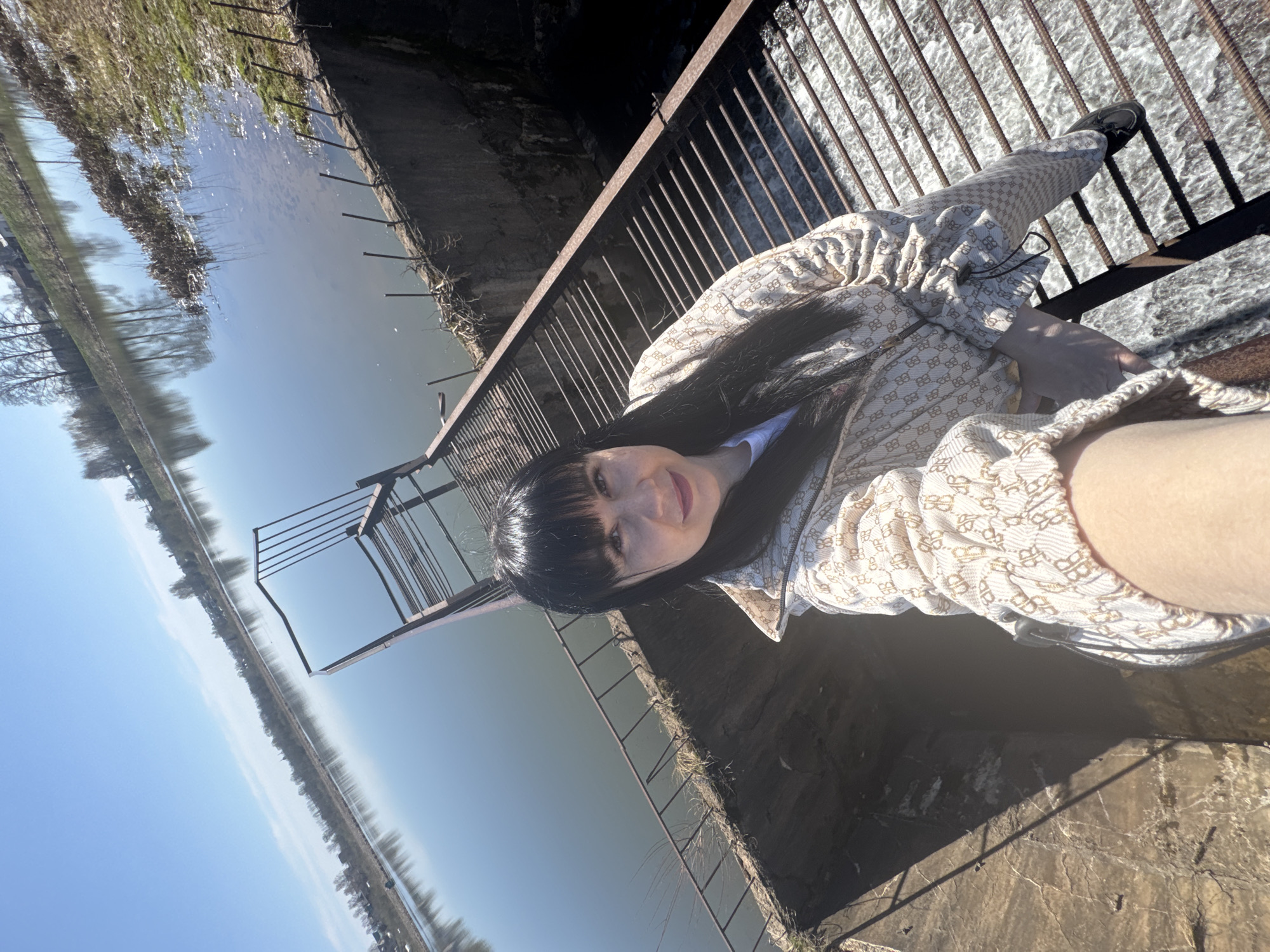 A medium-shot photograph captures a woman with dark hair and bangs standing on a bridge-like structure in Zav'yalovo, Russia. She is positioned to the right, wearing a light-colored patterned jacket and pants, with one leg extending towards the camera. The bridge has a metal railing.
To the left, a canal or river stretches into the distance, reflecting the blue sky. The environment is sunny, with vegetation visible along the banks. Additional metal structures and concrete edges suggest the location is an elevated crossing over water, potentially a small dam or a similar water control structure.