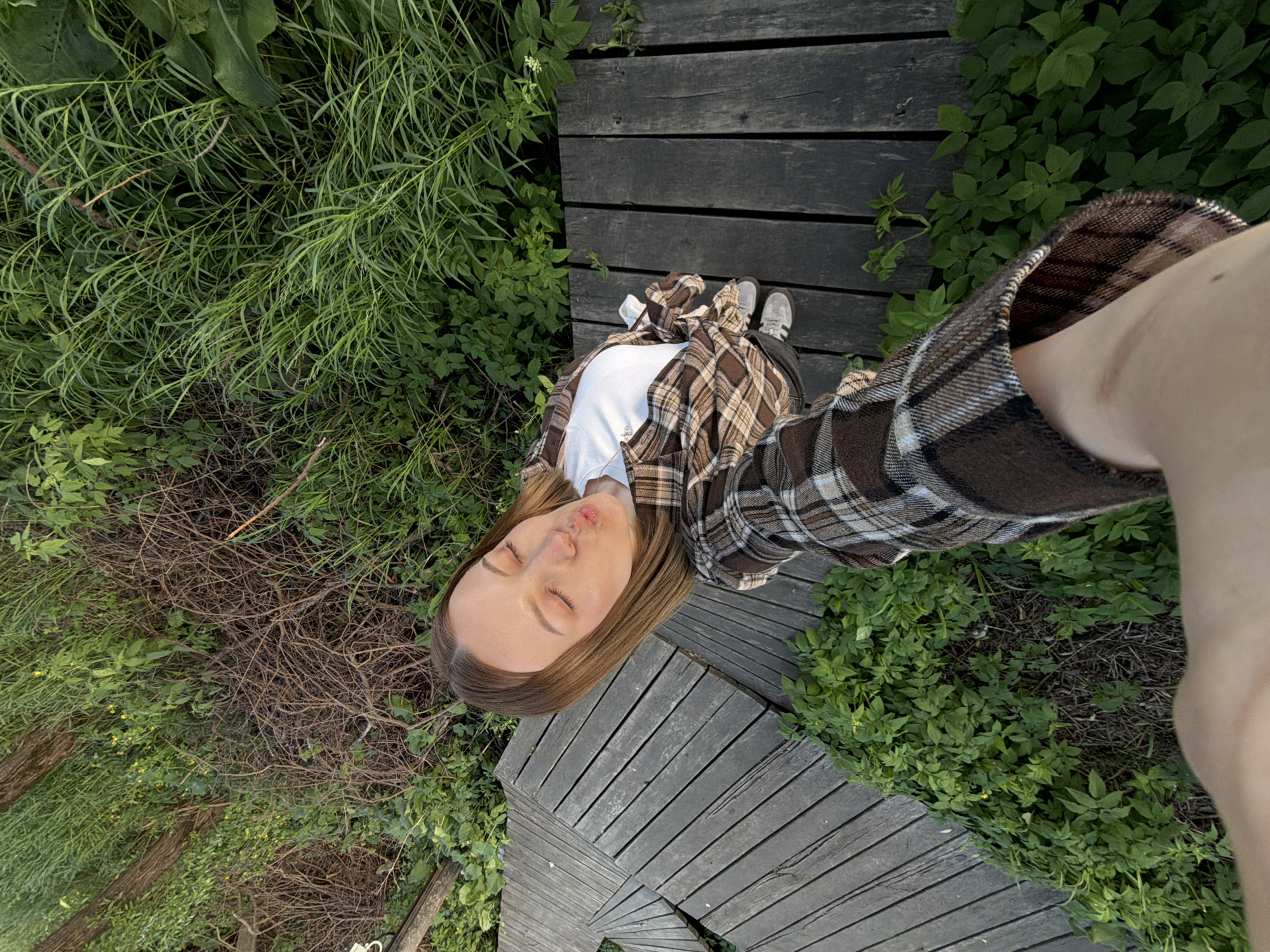 The image is a high-angle selfie taken on a wooden boardwalk in Sin'kovo, Russia. A young woman with light brown hair is the primary subject. She is wearing a white t-shirt and a brown and tan plaid flannel shirt. Her eyes are closed, and her lips are pursed. She stands on the boardwalk, which is surrounded by lush green vegetation and some dried branches. Her feet are visible, clad in grey sneakers. The camera angle is distorted, showing the person's arm extended to hold the camera. The environment is predominantly green, indicating a natural setting, possibly a park or wooded area. The boardwalk is made of dark-toned wood planks.